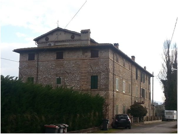 Edificio in mattoni con facciata a tre piani, sommità ad arco e persiane verde scuro, situato su una strada con un'auto e alberi.