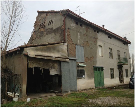 Edificio fatiscente a due piani con annesso capannone. Esterno in stucco grigio, con parti mancanti. Porta verde, alberi spogli nelle vicinanze.