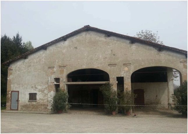 Vecchio fienile beige, usurato, con tre aperture ad arco, una piccola porta e un tetto marrone.