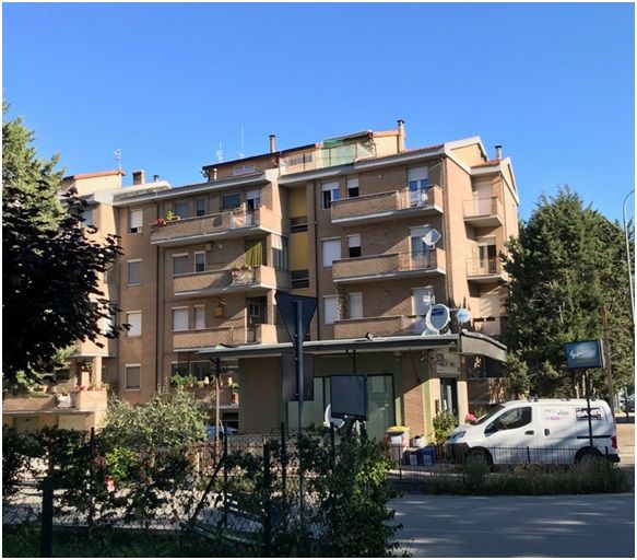 Condominio con balconi e un piccolo edificio con tenda da sole davanti; cielo azzurro.
