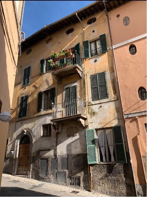 Edificio a più piani con vernice gialla scrostata, persiane verdi e un piccolo balcone in una strada stretta.