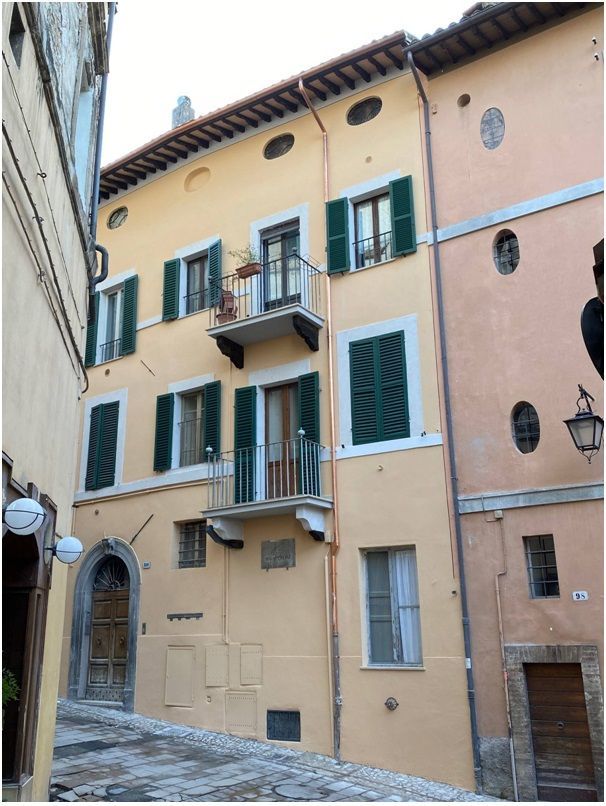 Edificio giallo di tre piani con persiane verdi, piccoli balconi e una porta di legno su una strada stretta.