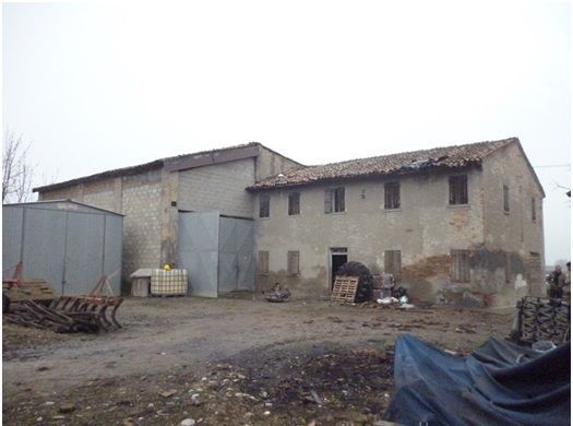 Vecchio edificio usurato dalle intemperie con tetto di tegole e capannoni di metallo in un ambiente rurale.