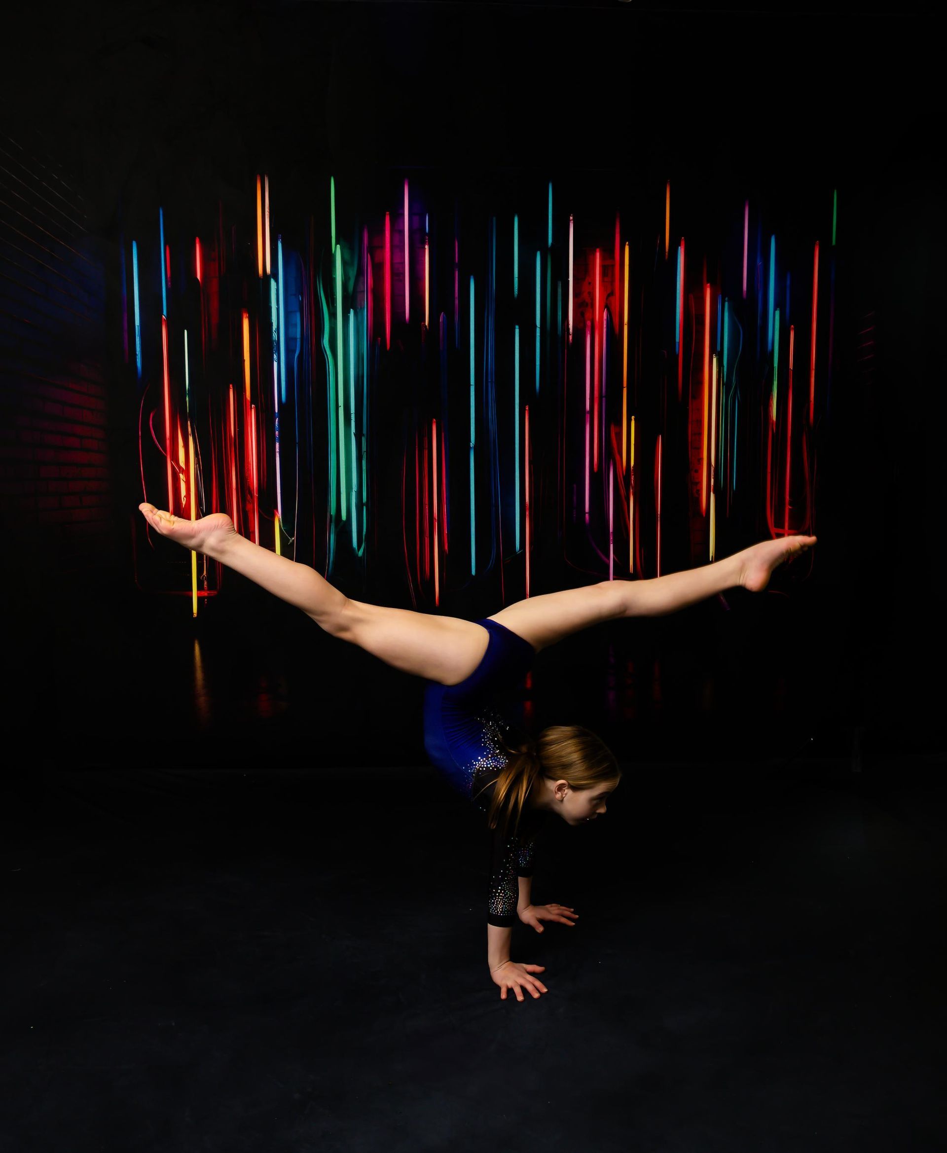 Gymnast in a handstand with legs split against a backdrop of colorful vertical lights.