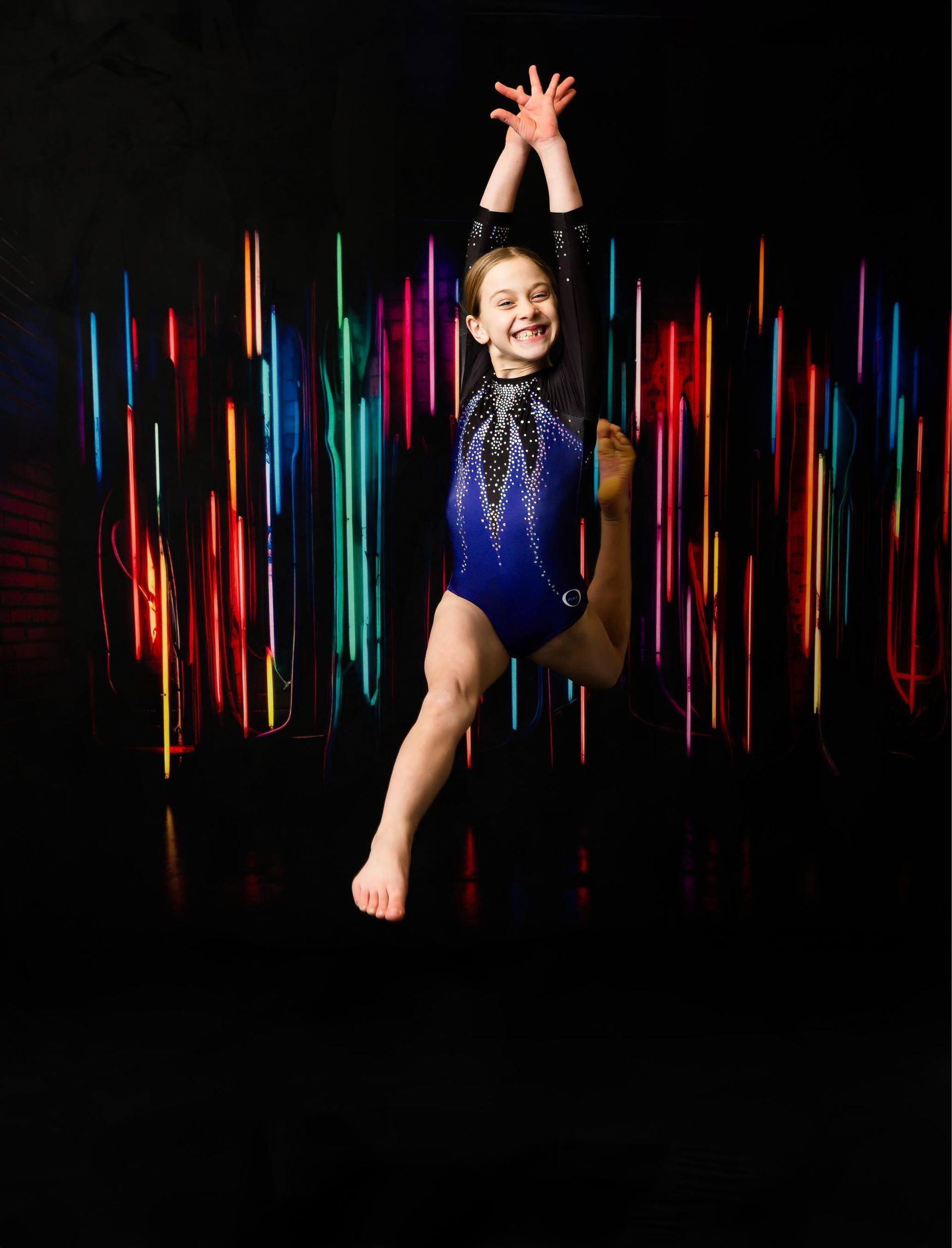 Gymnast in blue leotard jumping with one leg extended, arms raised. Bright, colorful light trails in background.