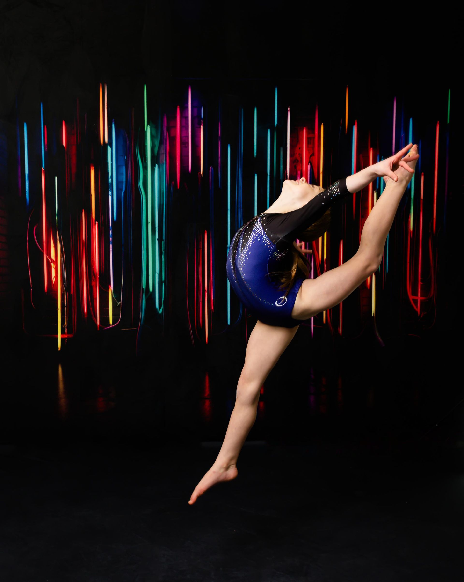 Gymnast in blue leotard leaps in a split, reaching for foot. Rainbow light backdrop.