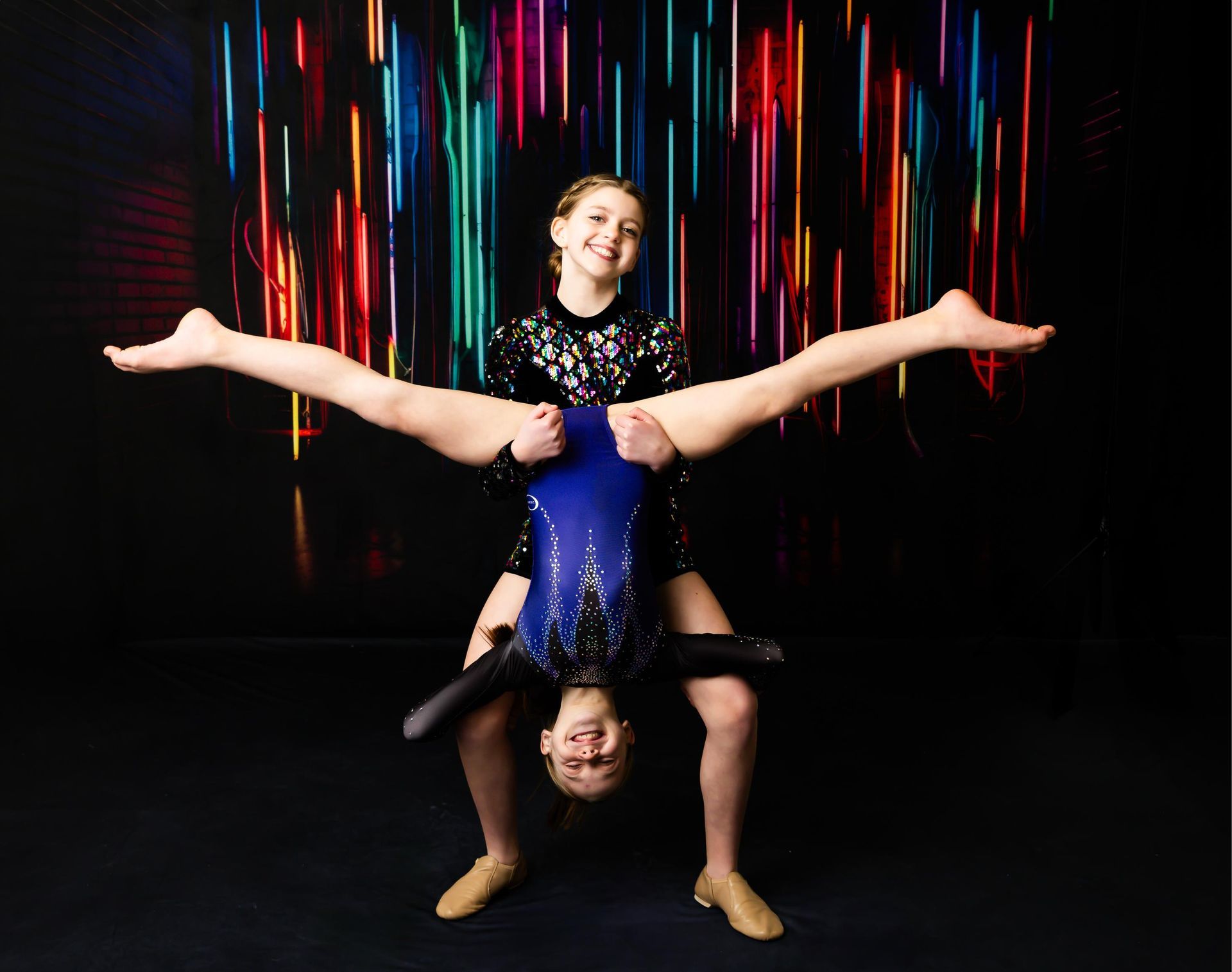 Two gymnasts in leotards performing a split and handstand against a colorful light backdrop.