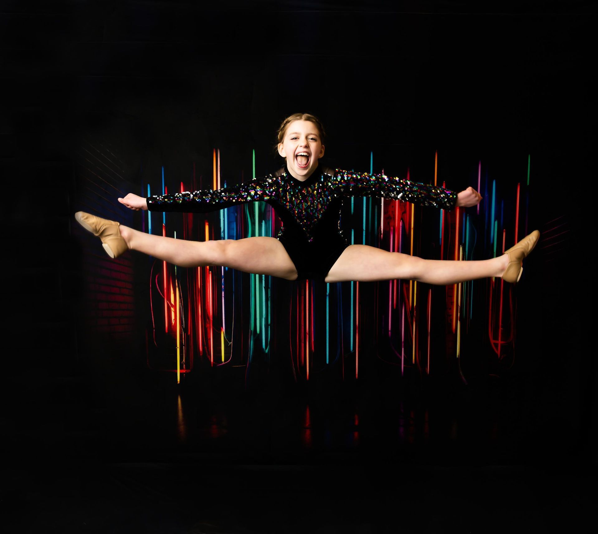 Girl in black leotard does the splits while jumping in front of colorful light display.