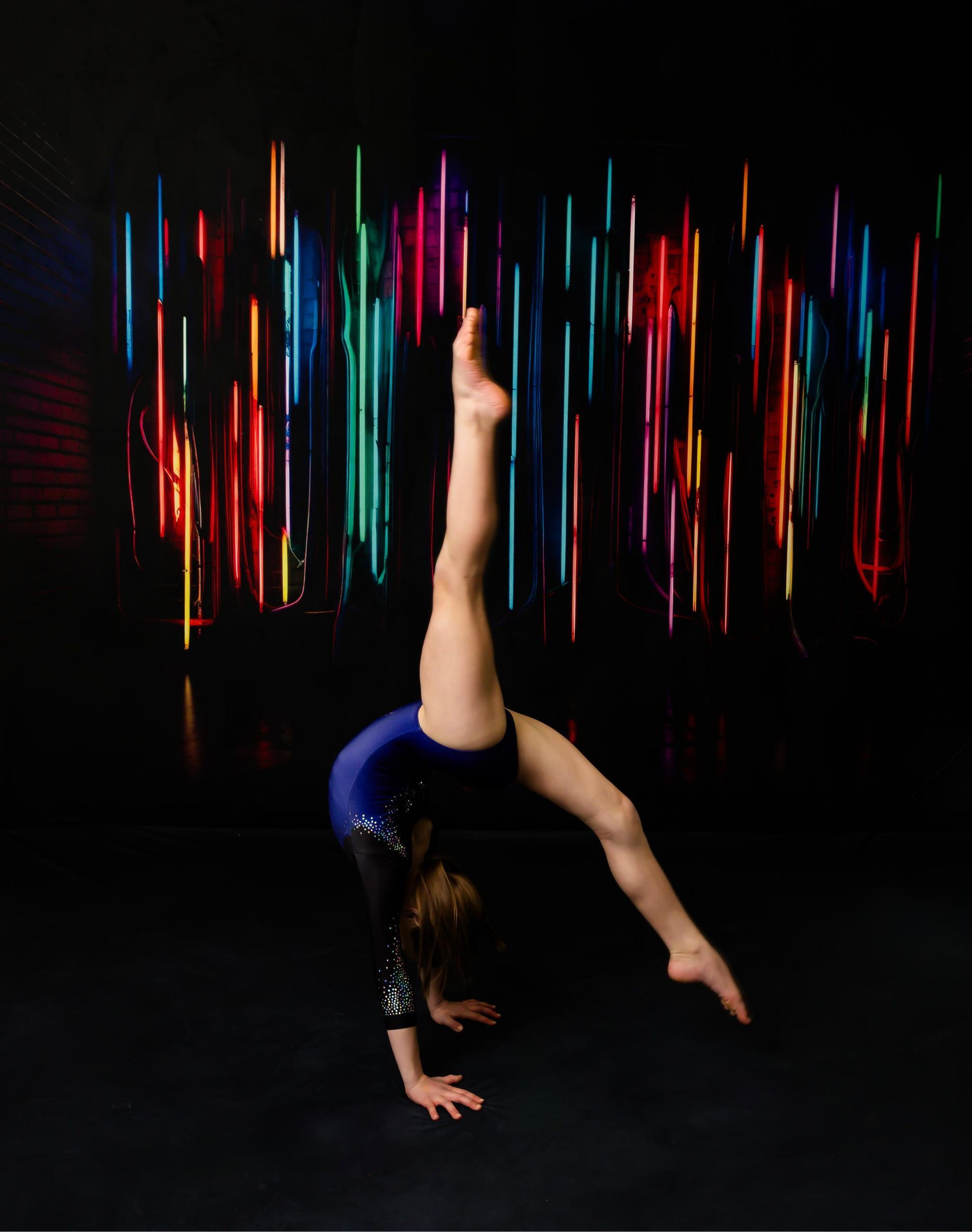 Gymnast in blue and black leotard performs a handstand with one leg extended high against a neon light backdrop.