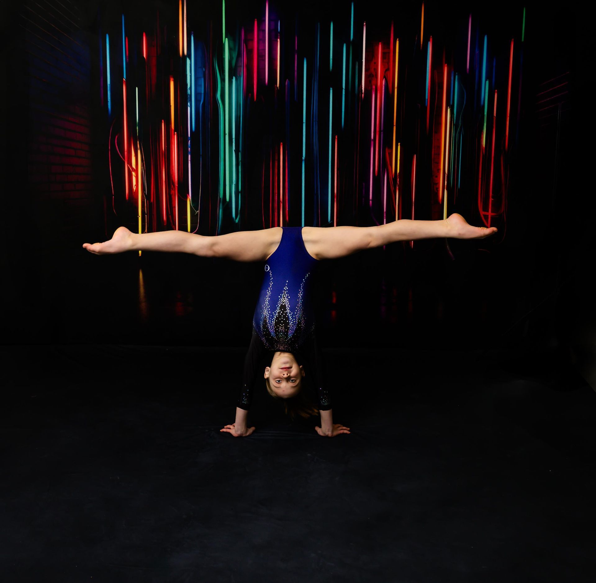 Gymnast in blue leotard performs handstand with legs split wide, colorful light streaks in background.