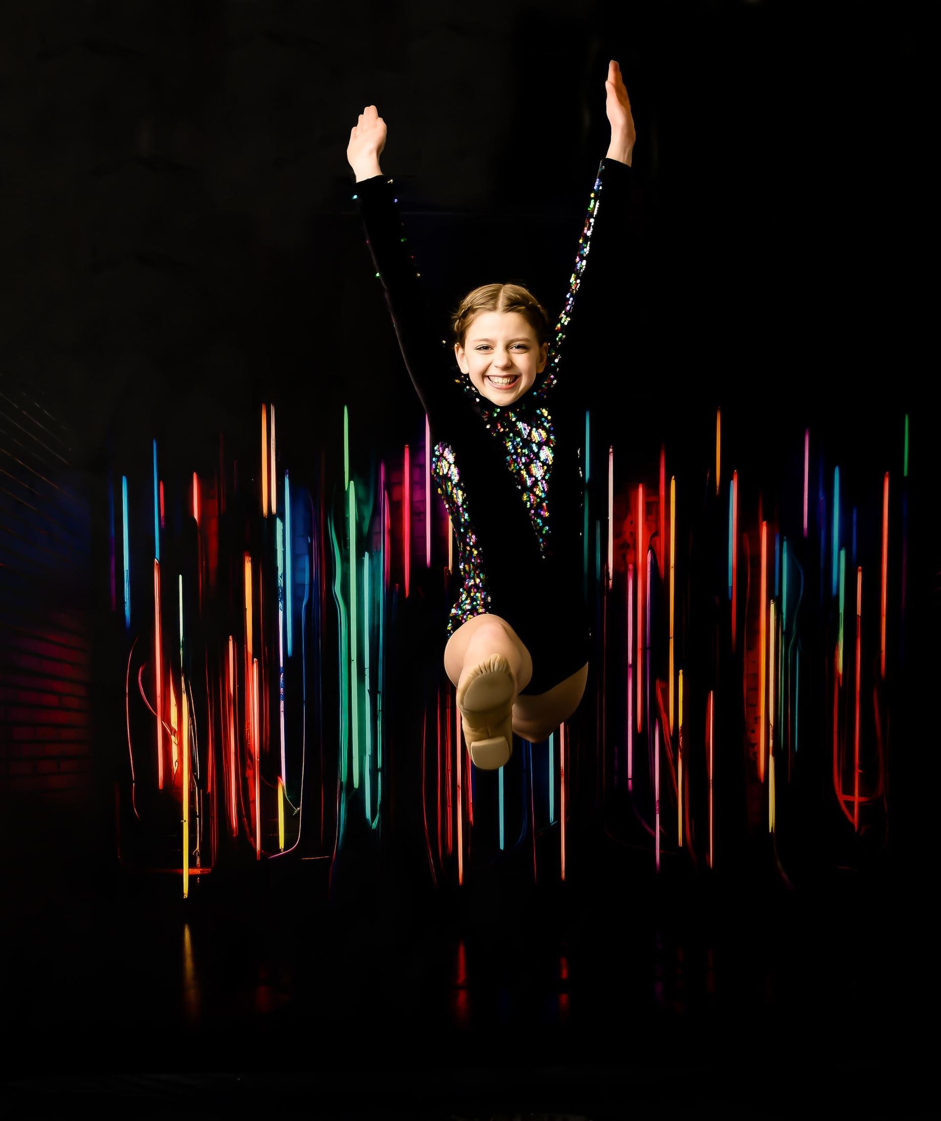 Girl leaps in the air, arms raised, smiling. Black outfit, colorful background lights.