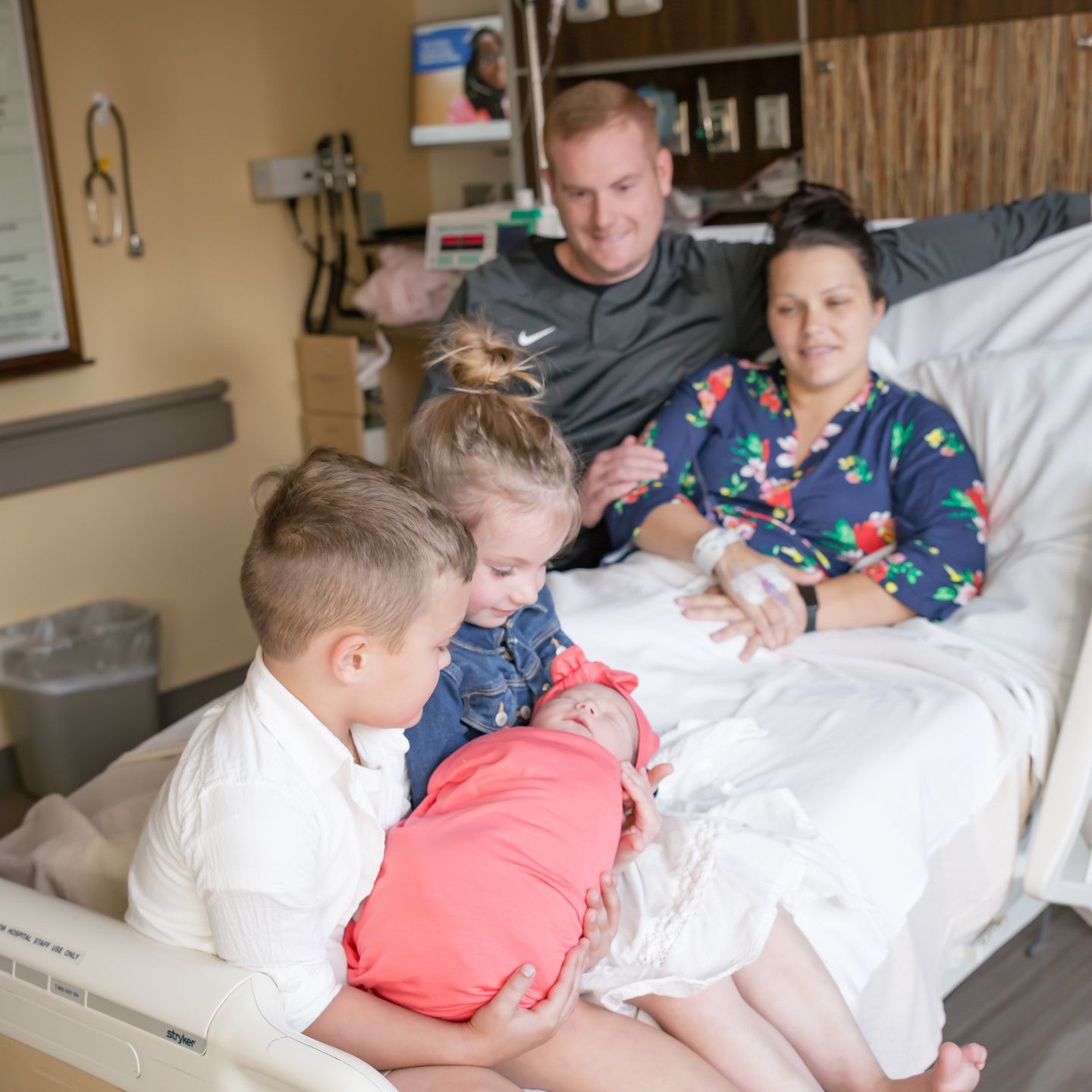 Family of five in a hospital room, mother in bed holding newborn, siblings looking on, father smiling.