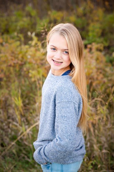 Girl with long blonde hair and braces smiles, standing in field, wearing blue sweater and jeans.