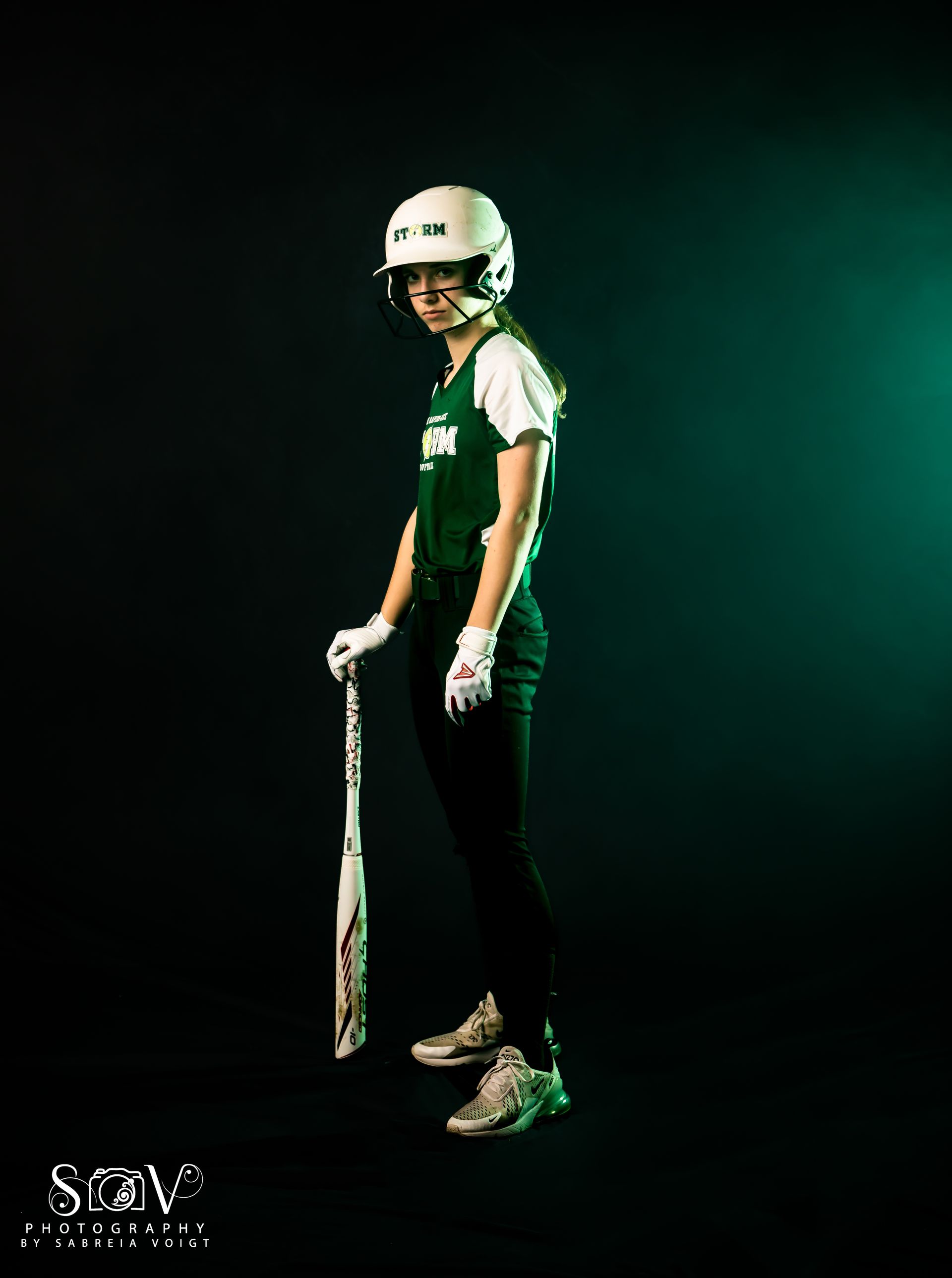 Softball player in green uniform and helmet, holding a bat. Dark setting, green light.