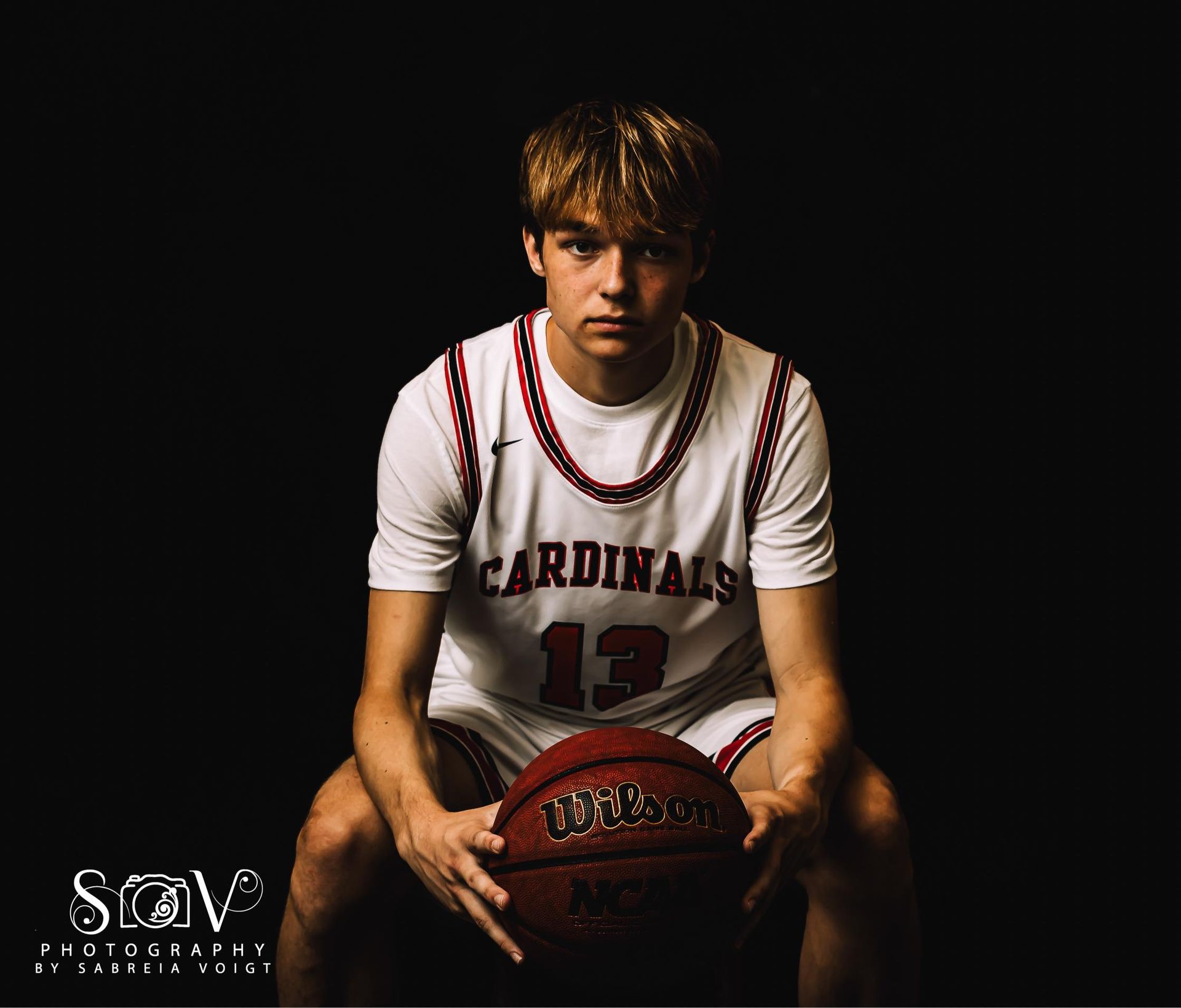 Basketball player in a Cardinals jersey, number 13, holding a ball, sitting. Black background.
