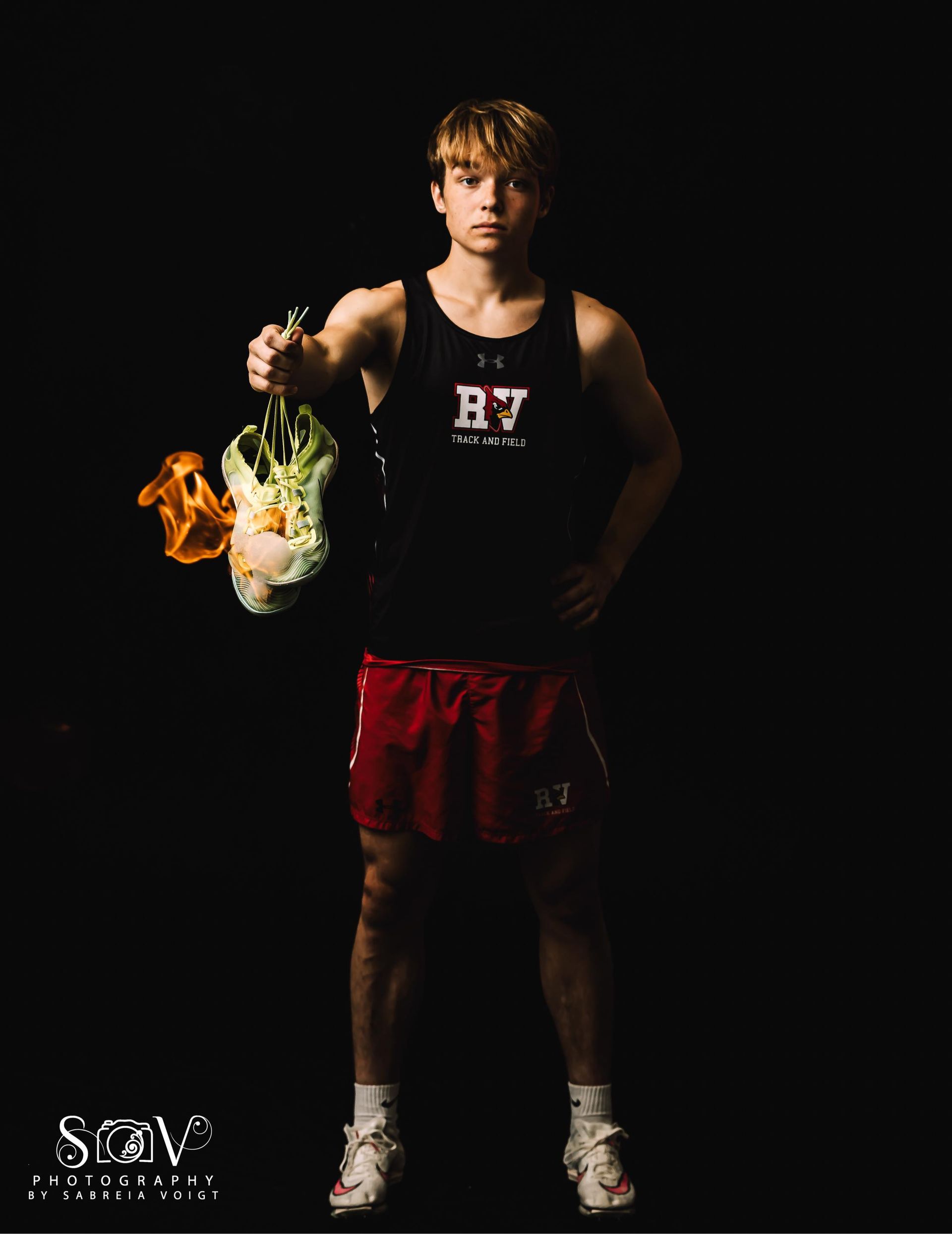 Athlete holding burning shoe, wearing black tank top, red shorts, white shoes, standing in a dark setting.