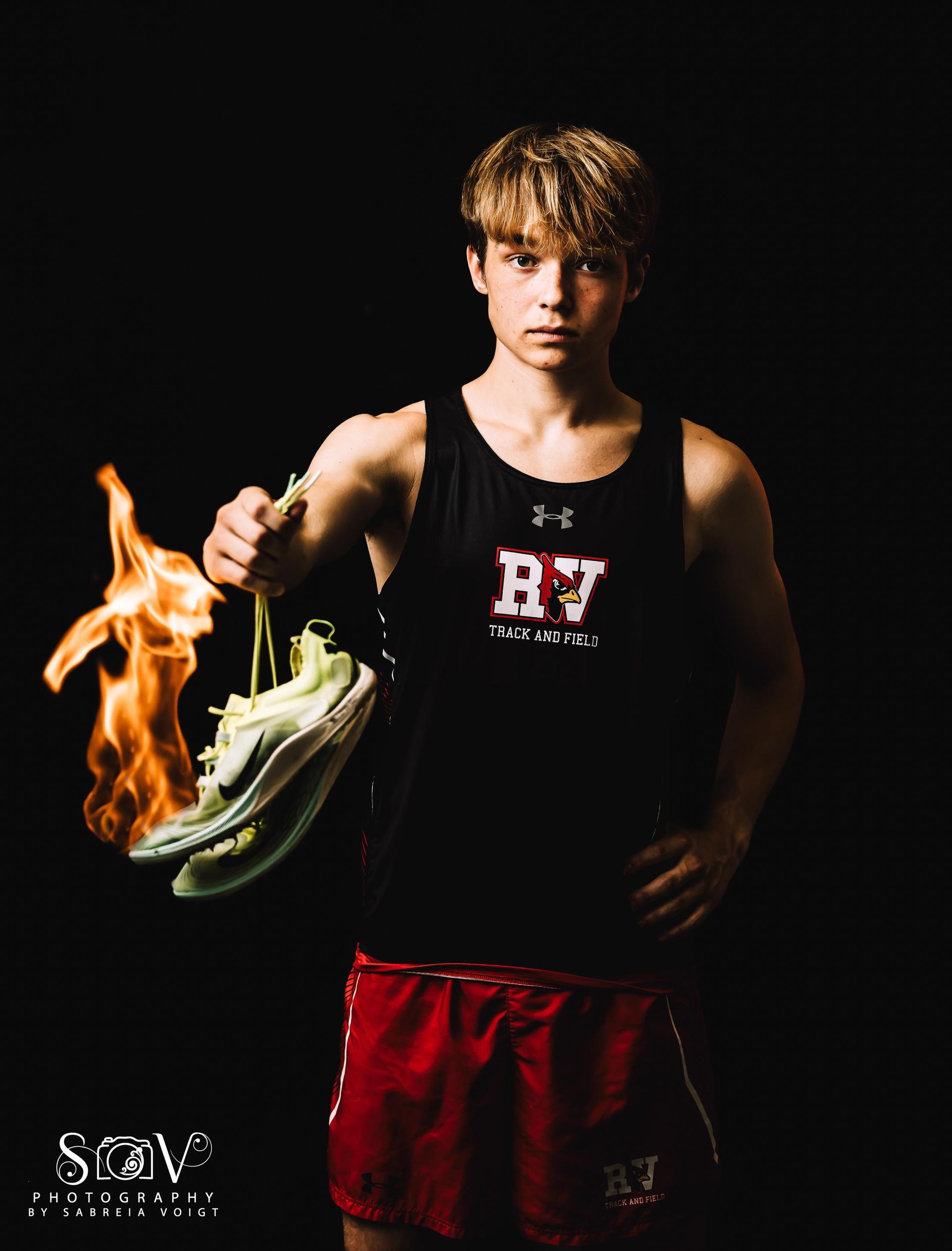 Runner in black singlet and red shorts holds flaming shoe against black background.