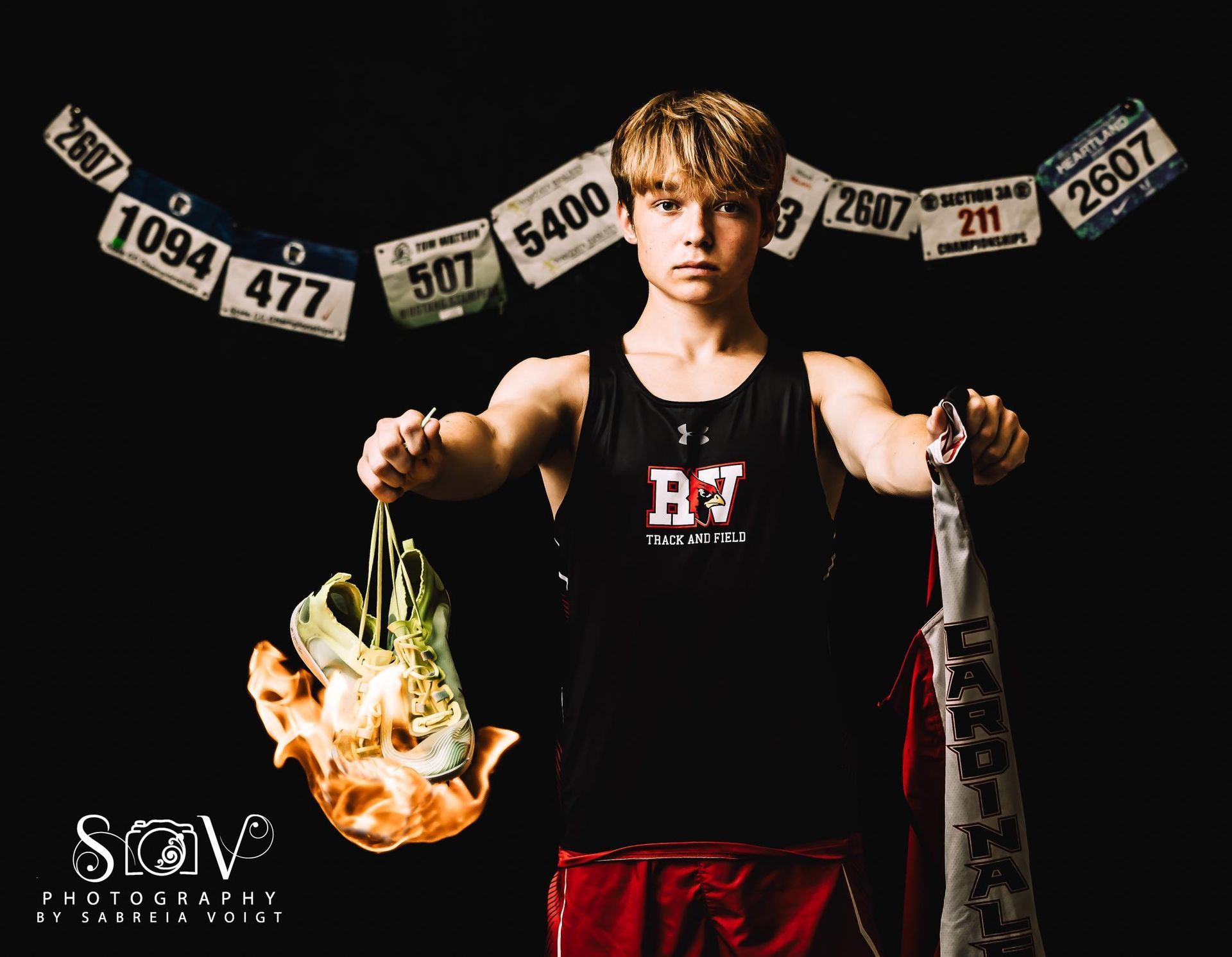 Teen athlete holding shoes and towel, bibs overhead. Black tank top, red shorts, black background.