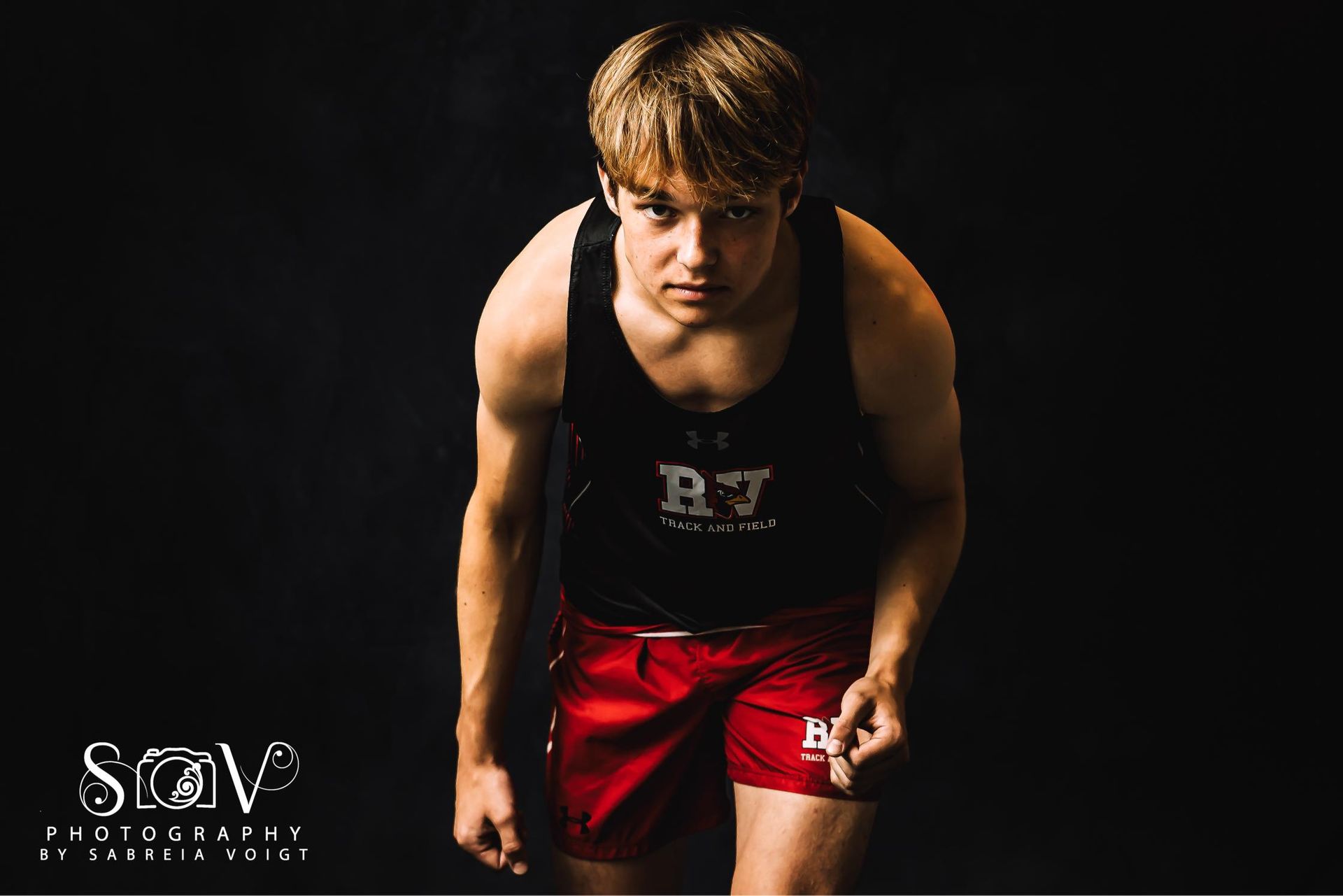 Wrestler in black tank top and red shorts, stance ready. Dark background.
