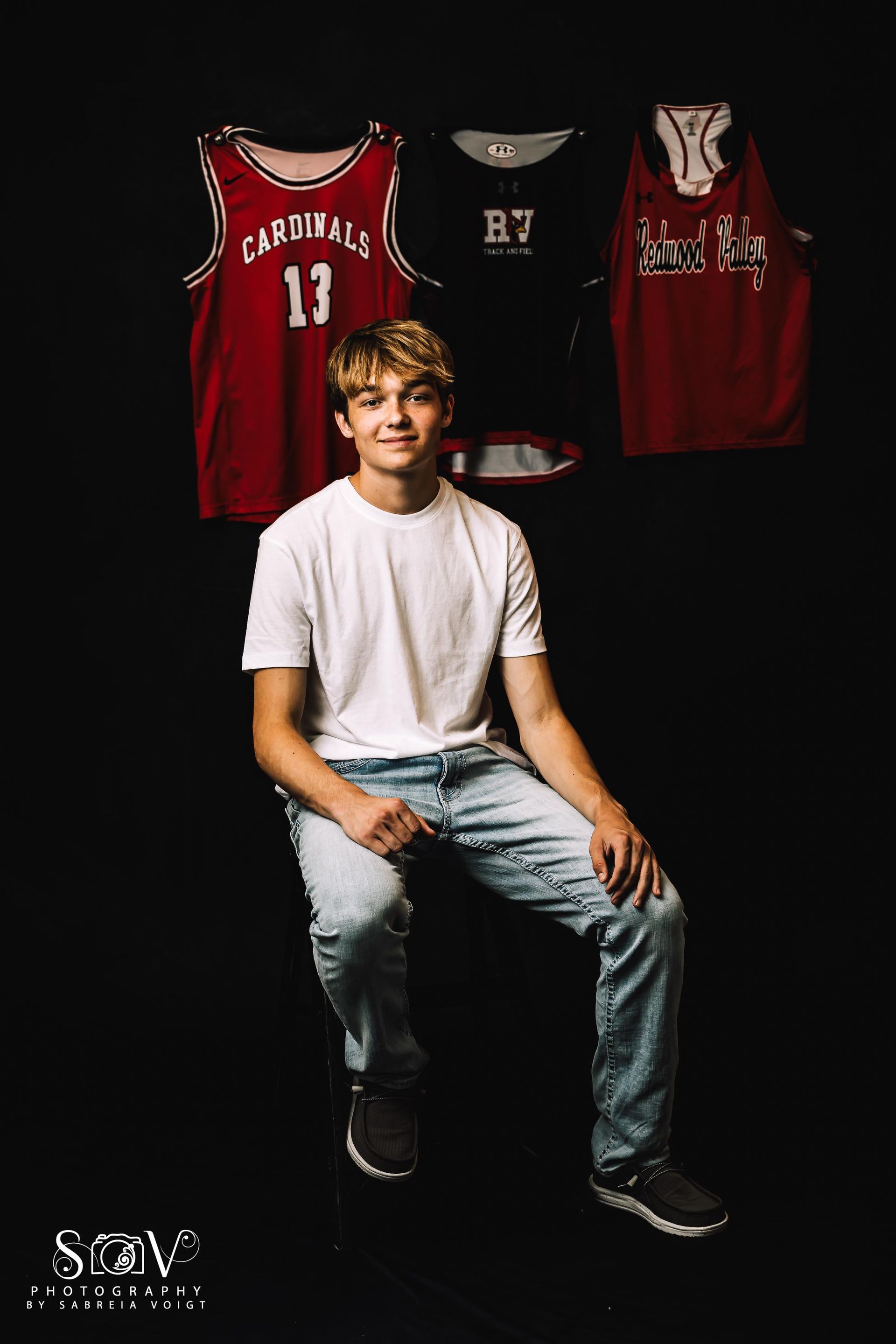 Teenage boy seated, smiling, in front of jerseys. Black background, wearing white shirt and jeans.