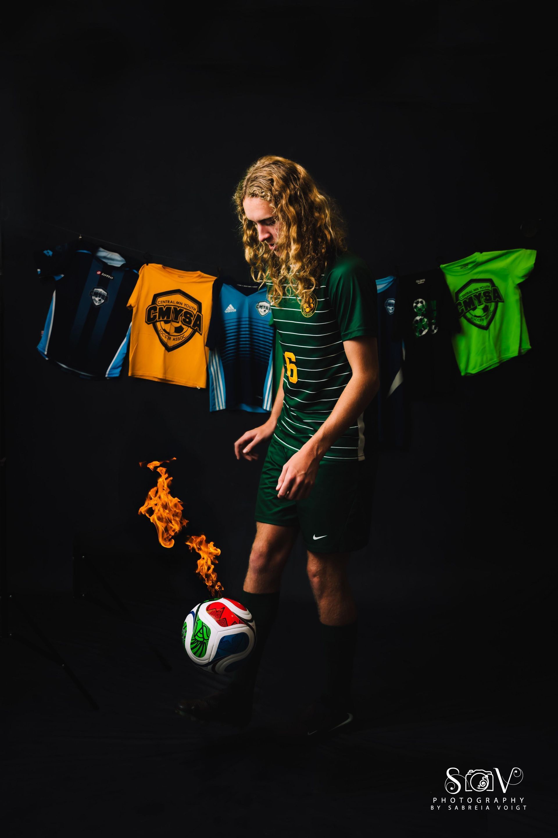 Soccer player in green uniform, juggling a flaming ball, surrounded by jerseys. Black backdrop.