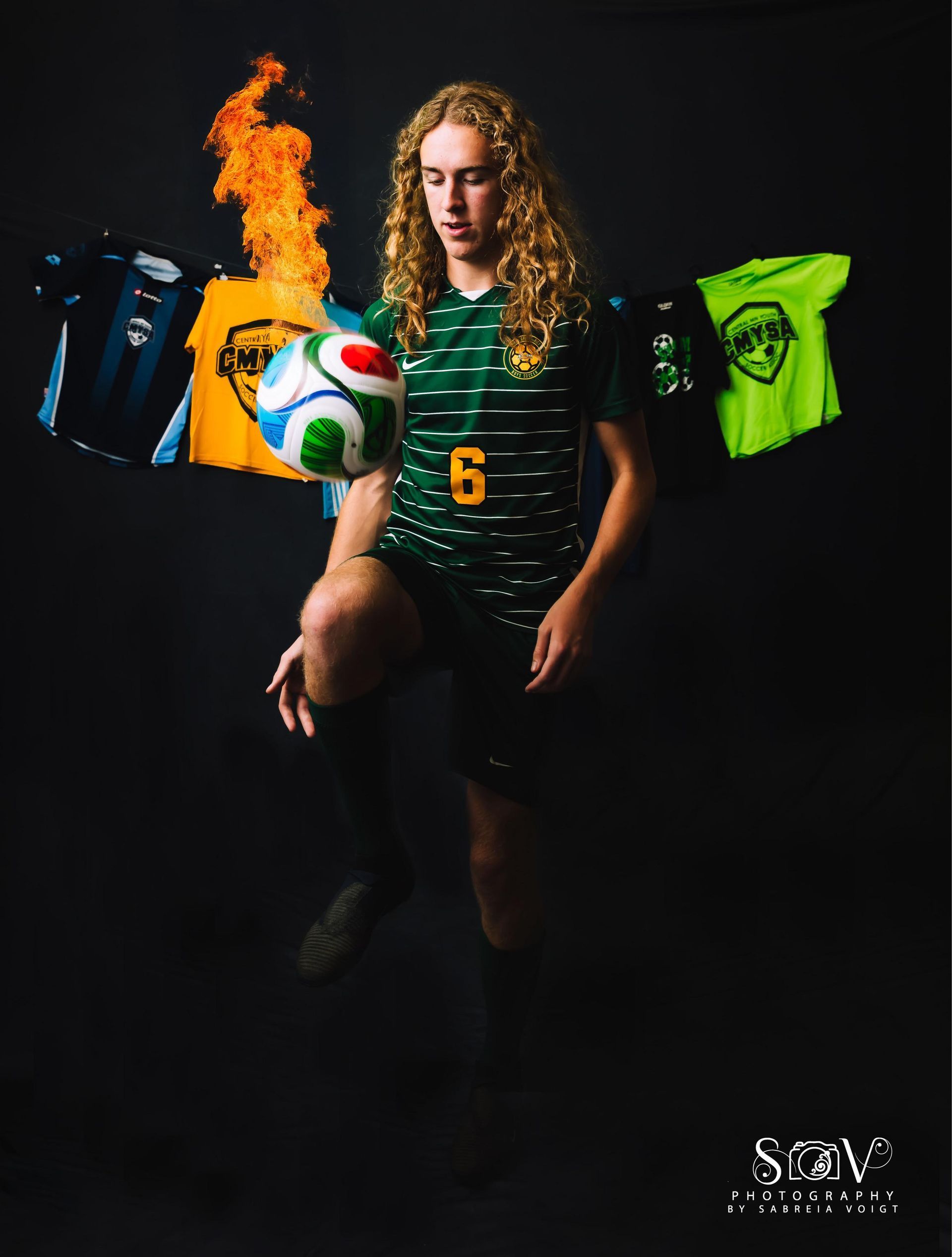 Soccer player balances ball on his knee, wearing green jersey with flaming backdrop and jerseys.
