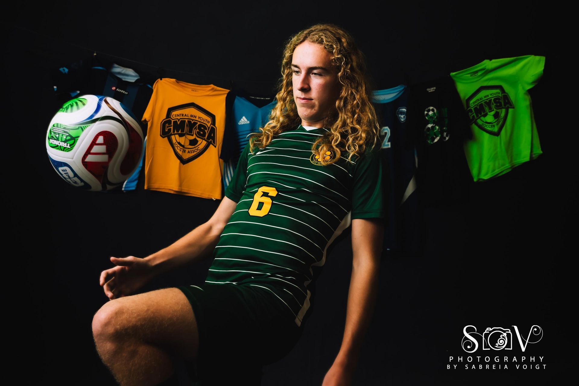 Soccer player in green jersey with number 6, posing with ball. Soccer shirts and ball in background.