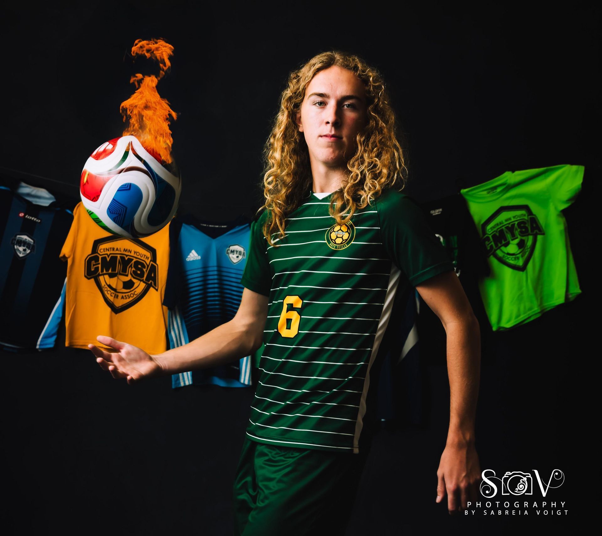 Soccer player in green jersey with number 6, holding flaming soccer ball, jerseys in background.