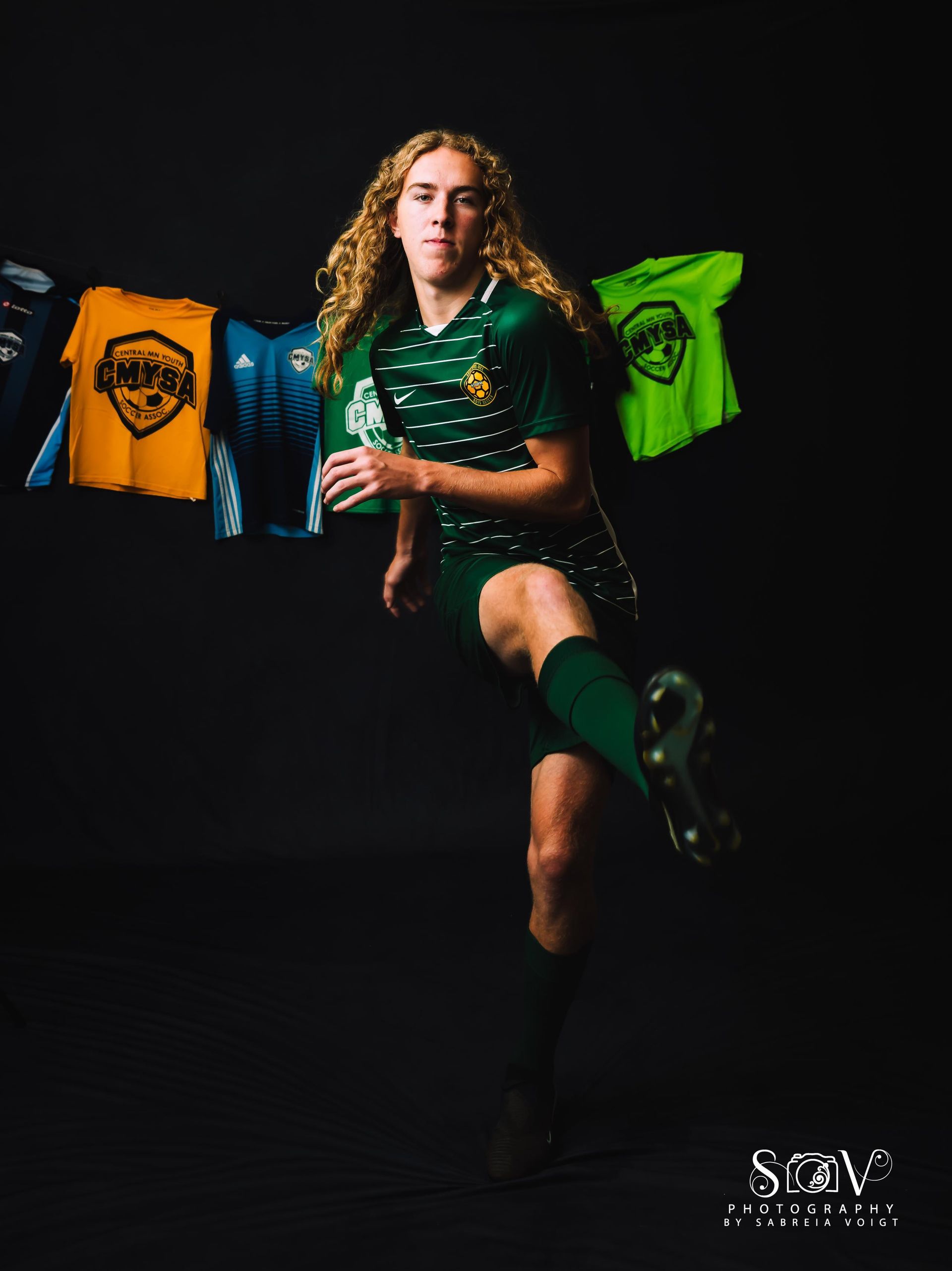 Soccer player in green uniform kicking, dark background with jerseys.