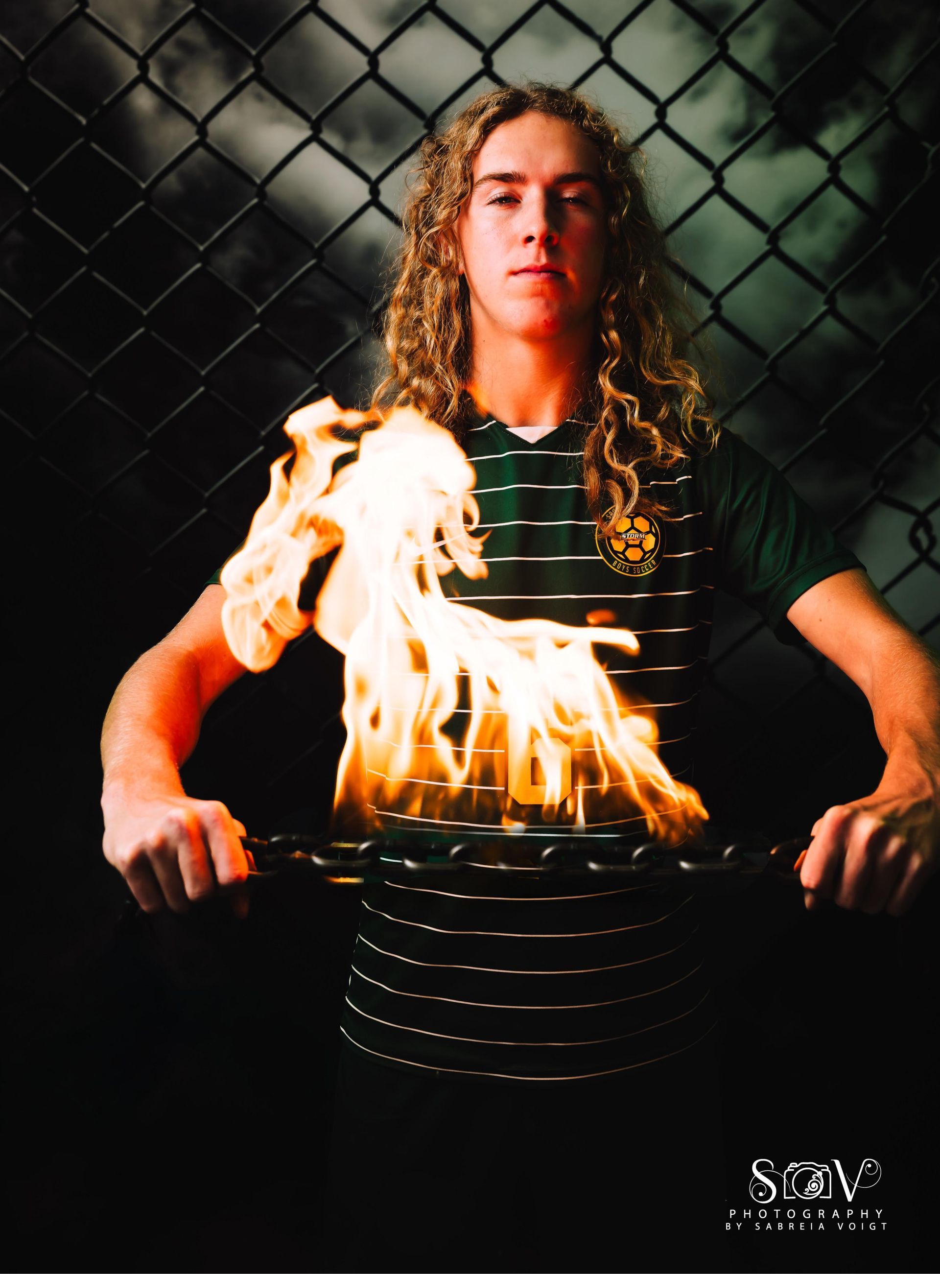Man with curly hair holds chain engulfed in flames. Wearing a green and white striped shirt. Set against a chain link fence.
