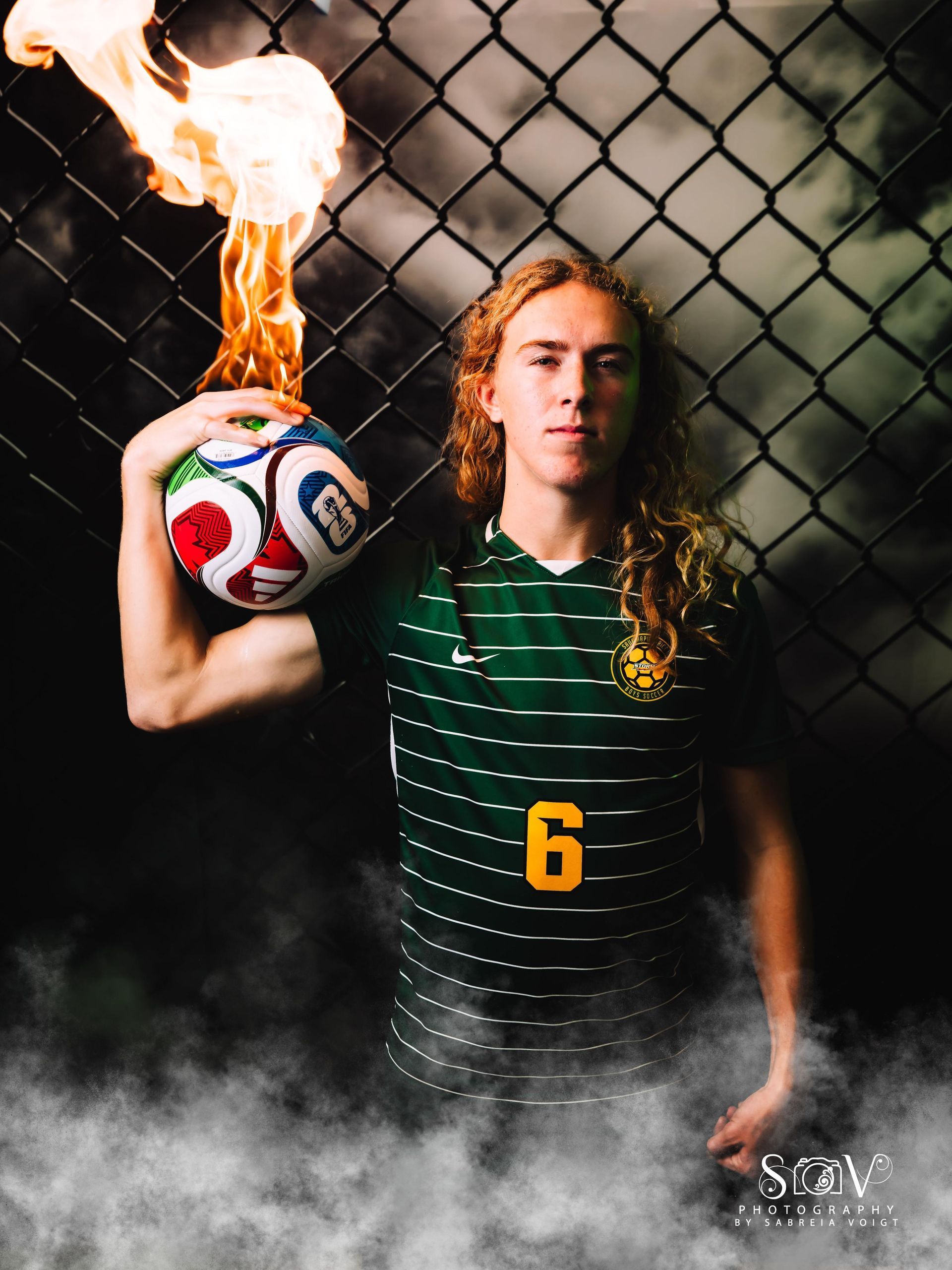 Soccer player holding a flaming ball in front of a chain-link fence. They wear a green and gold jersey with the number 6.