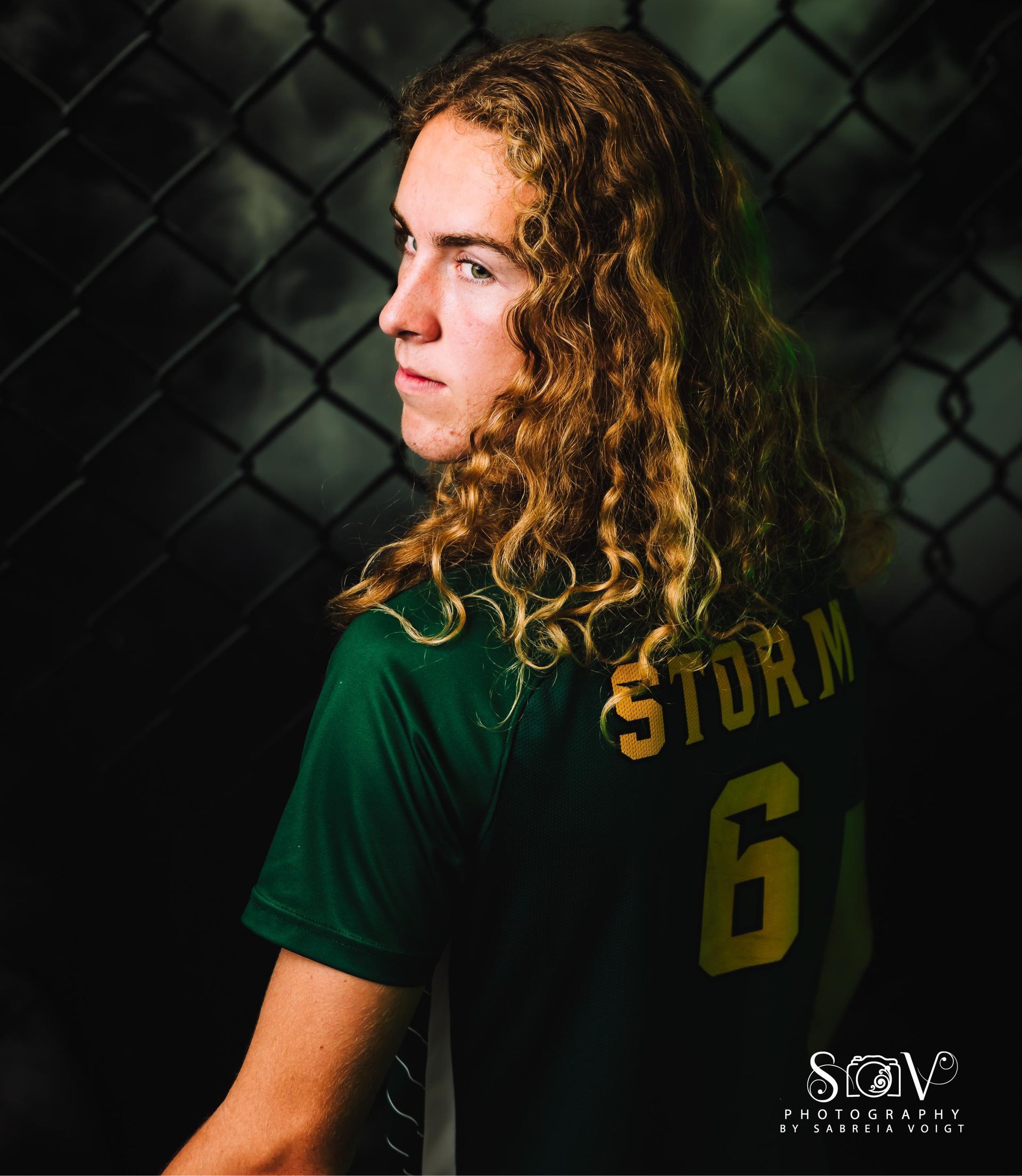 Person with long curly hair wearing a green and yellow jersey, turned looking back; fence background.