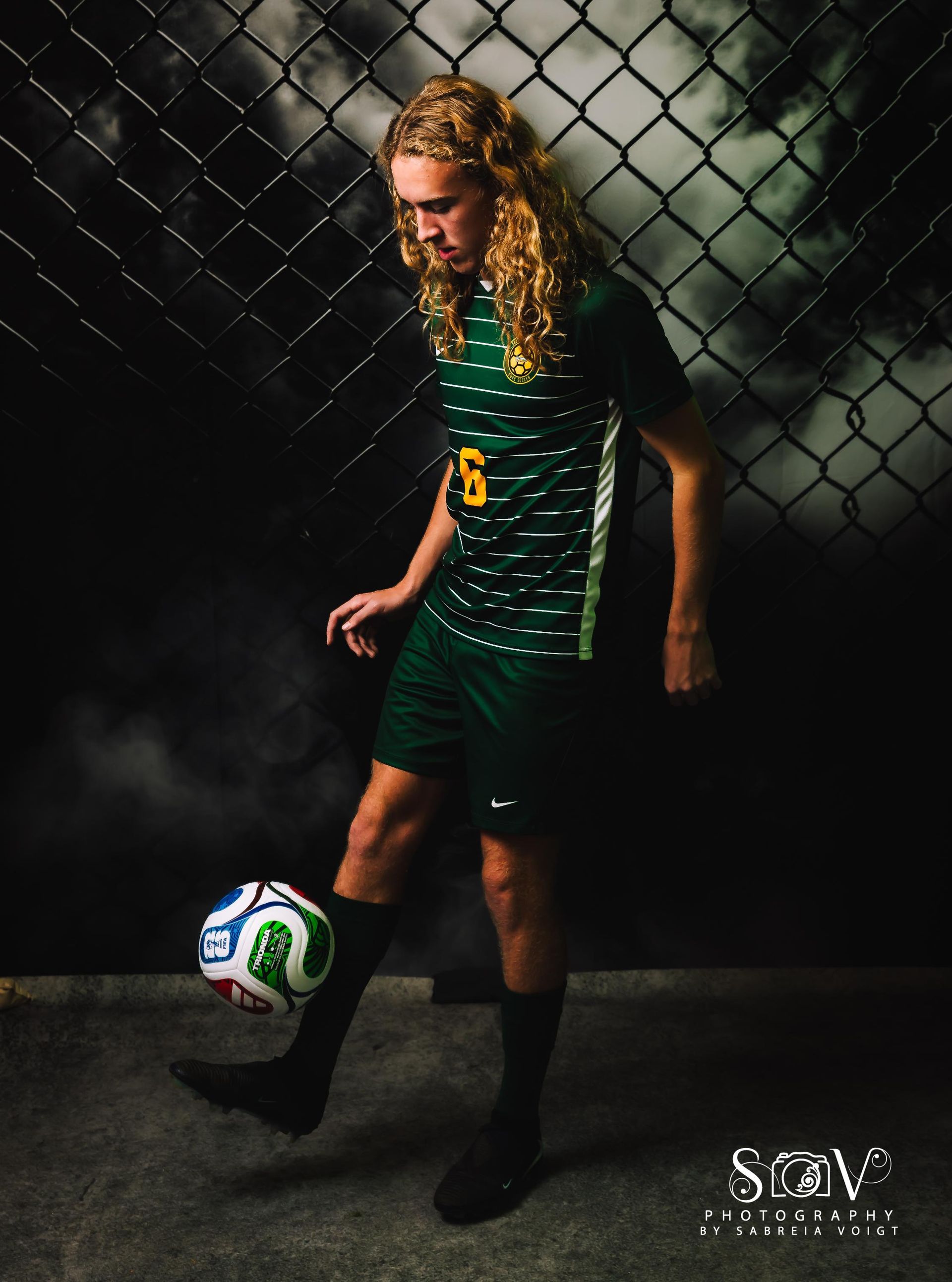Soccer player in green and white uniform juggling a ball, standing against chain link fence.