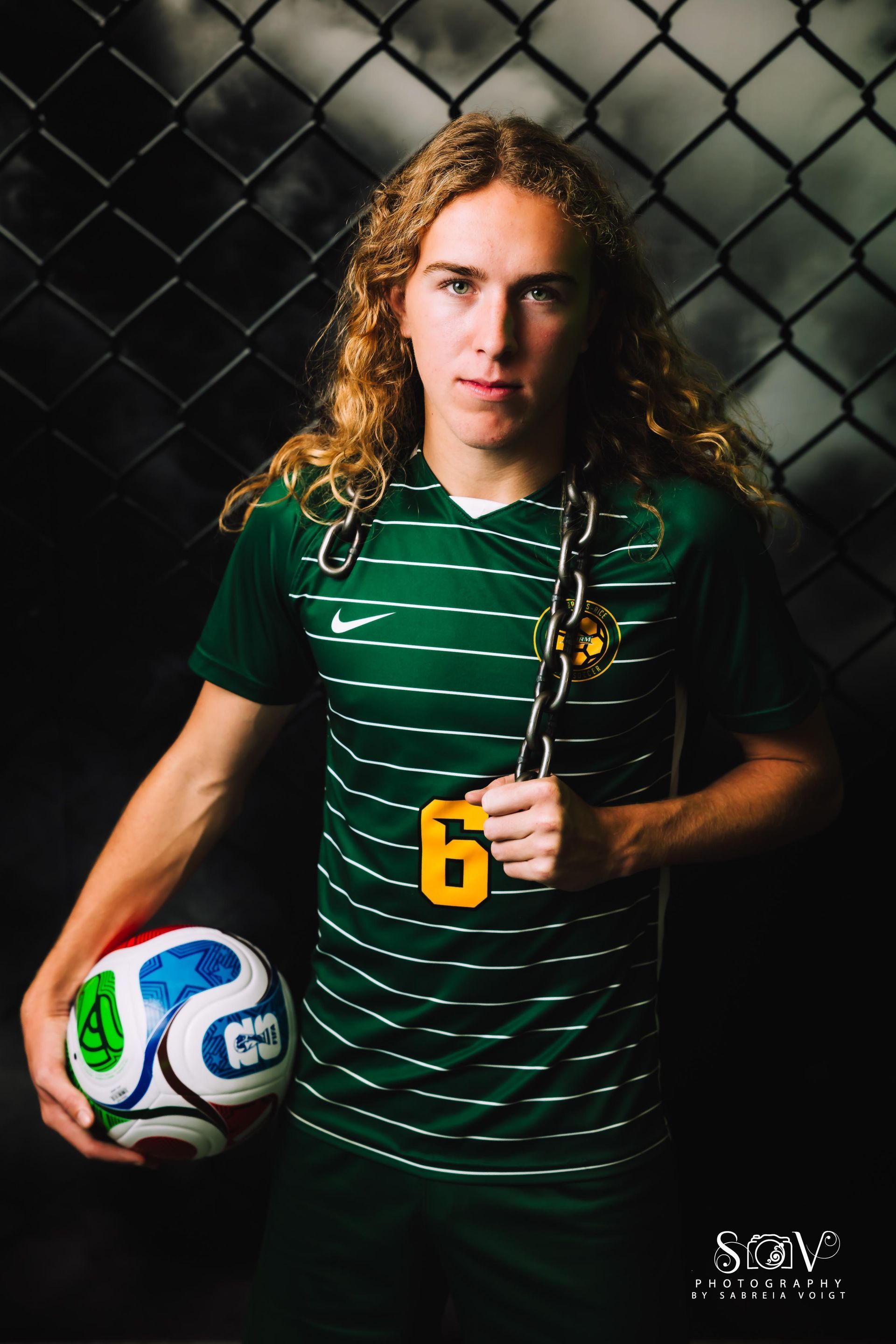 Soccer player in green jersey with long hair, holding ball and chain, standing in front of a chain-link fence.