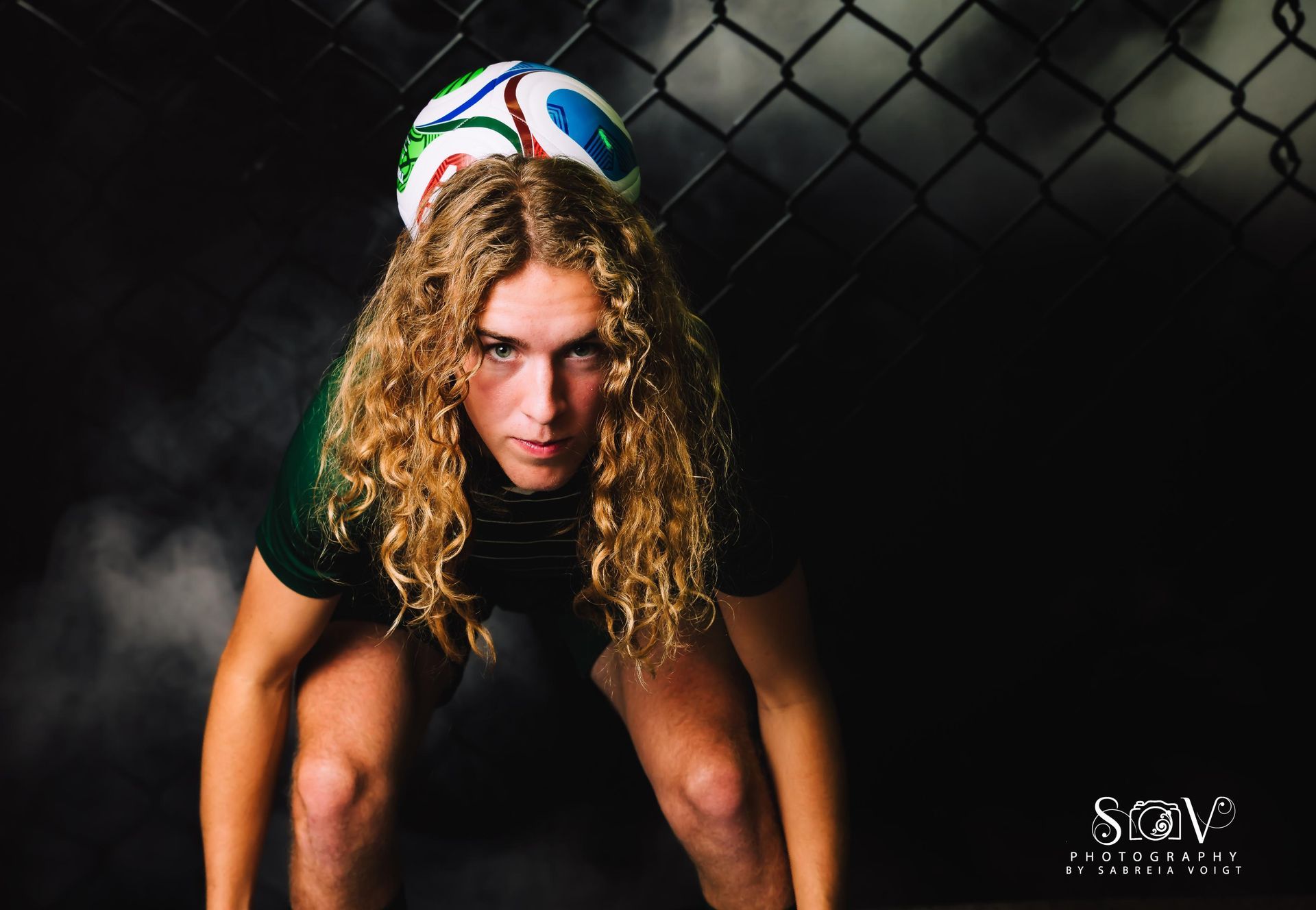 Person balances soccer ball on head, bent over in a dark setting, chain link fence in background.