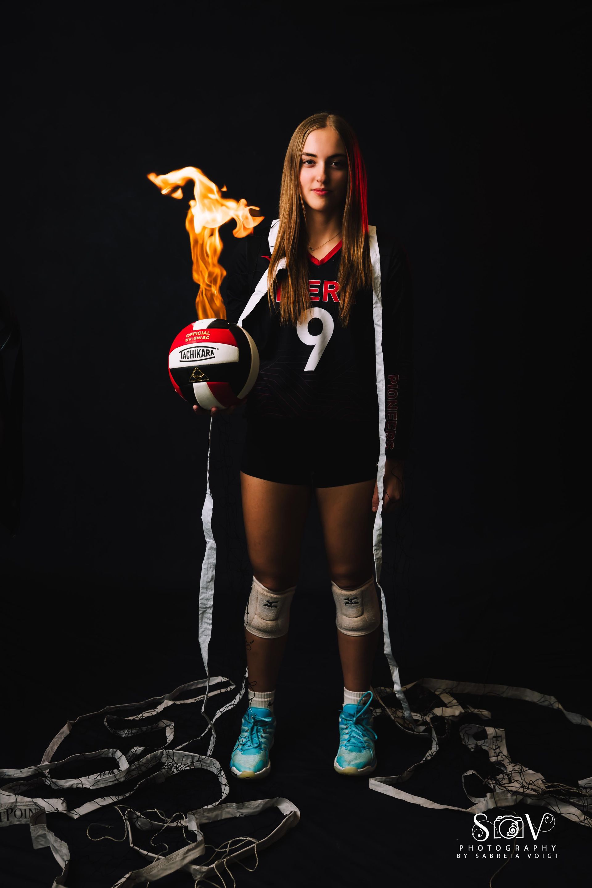 Volleyball player holding flaming ball, wearing uniform and knee pads, surrounded by shoes, black background.