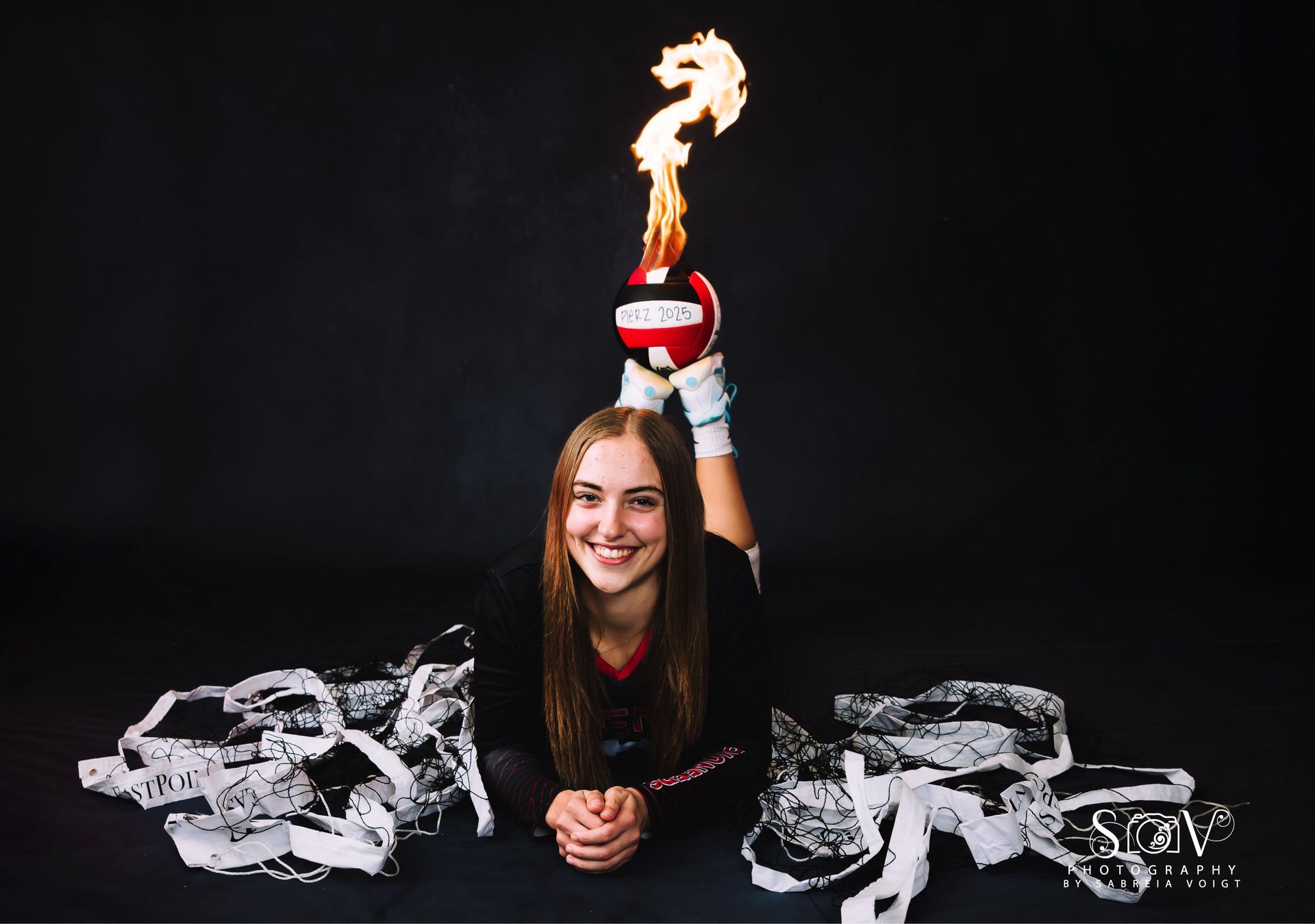 Woman holding a flaming volleyball, smiling. Black background, scattered gear.