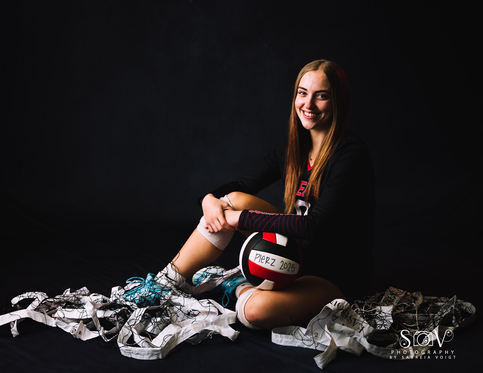 Young woman with volleyball, smiles while sitting on floor with uniforms. Black background.