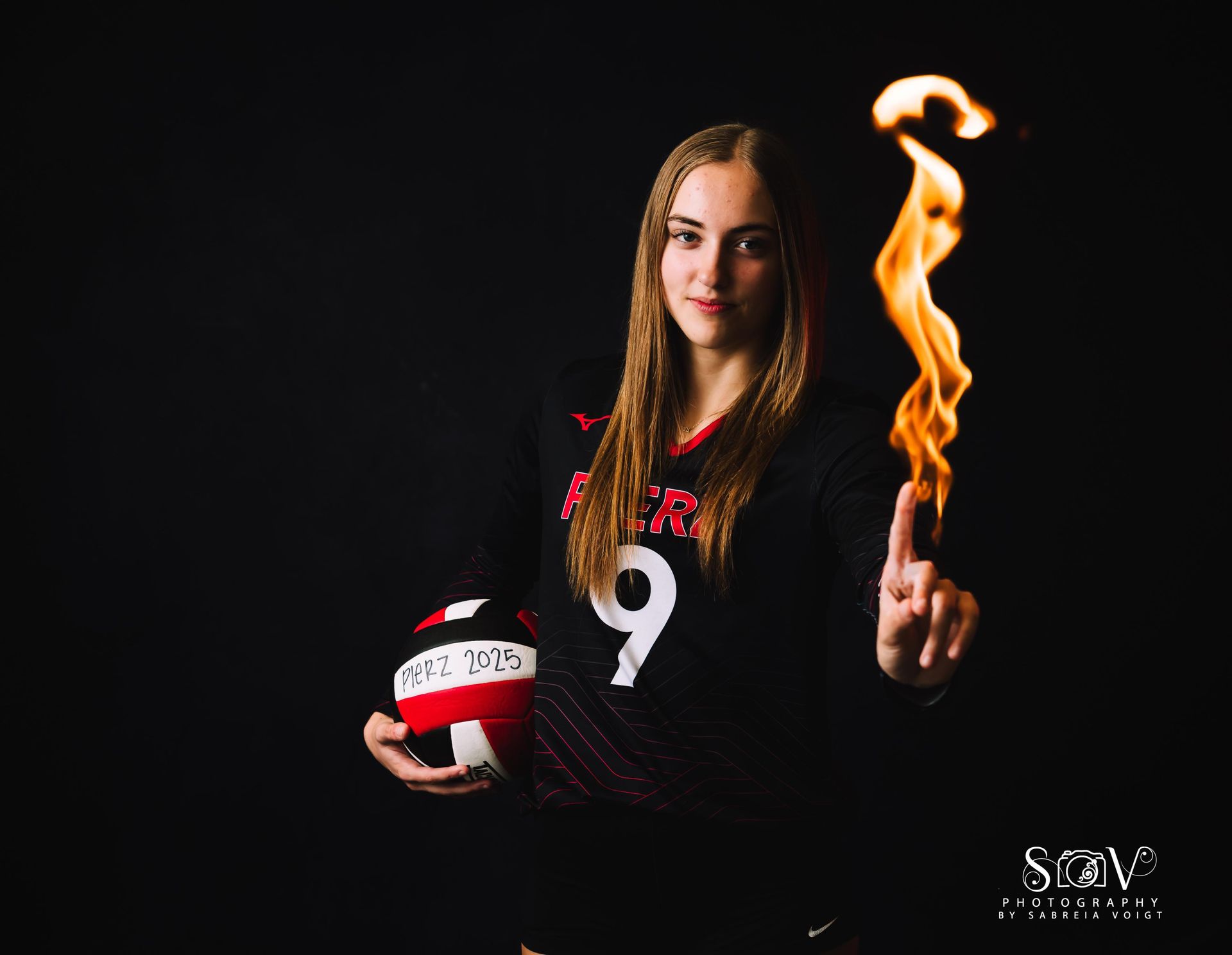 Volleyball player in black jersey holds ball, finger with flame against black background.