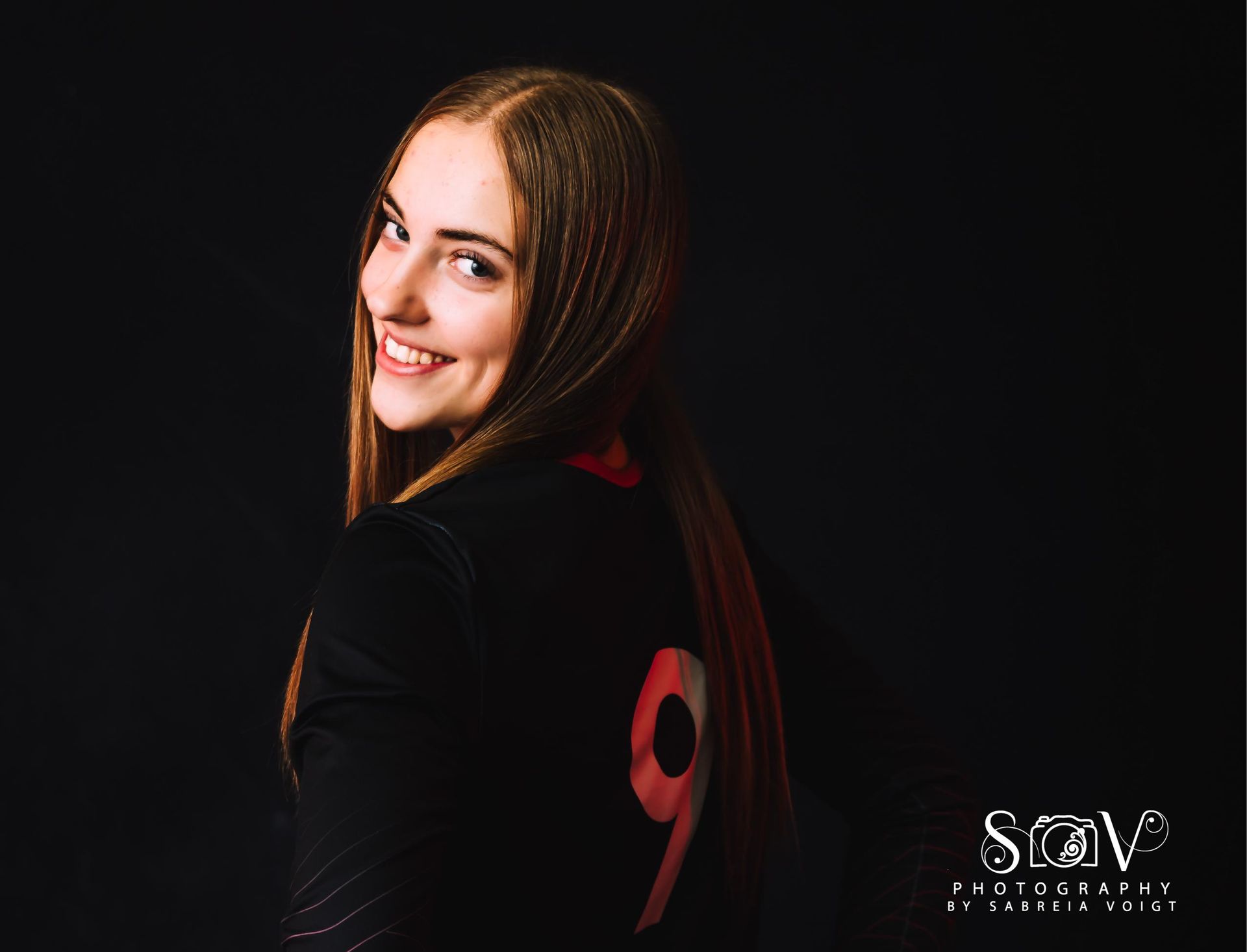 Young person with long hair smiles, looking over shoulder. Black shirt with large red 