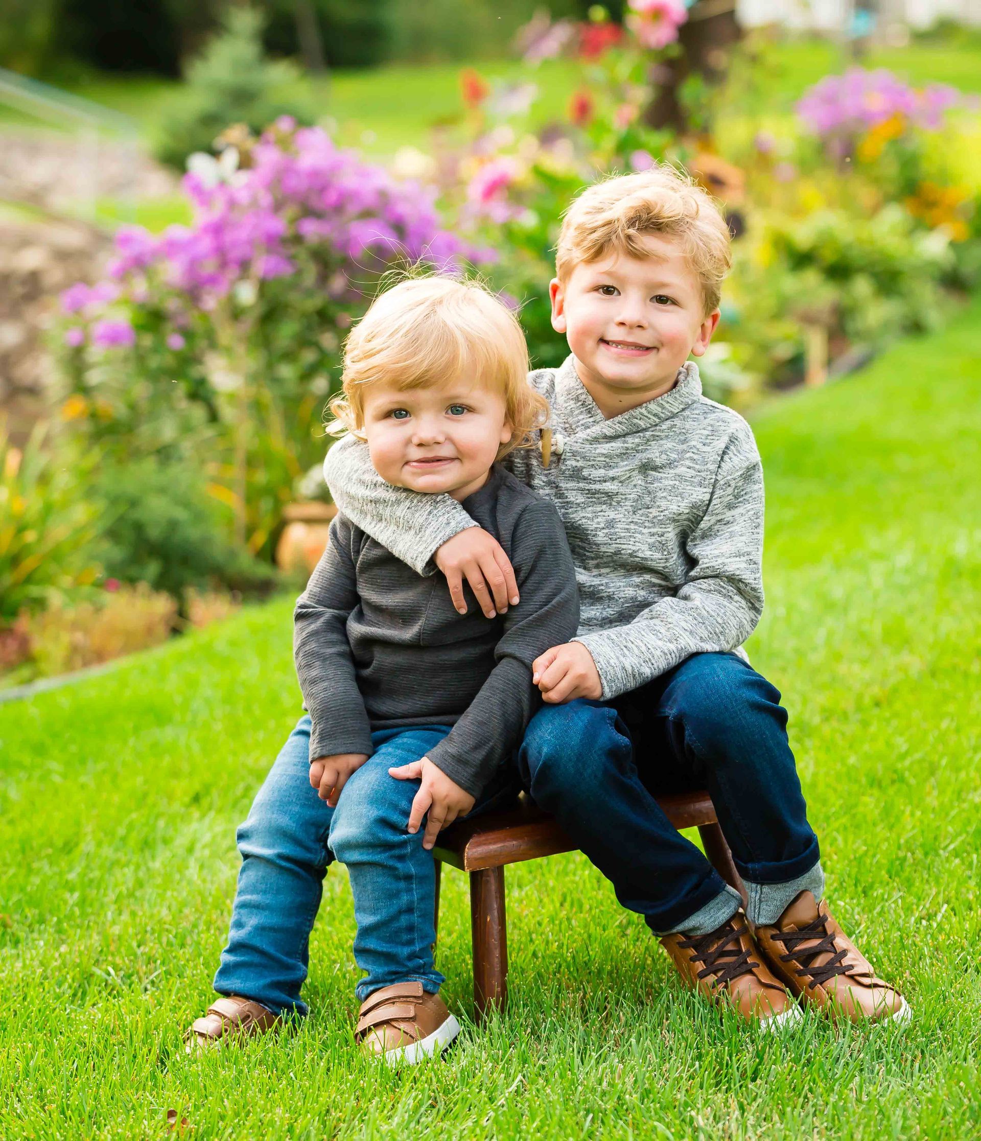Two young boys in a garden, one with an arm around the other, smiling.