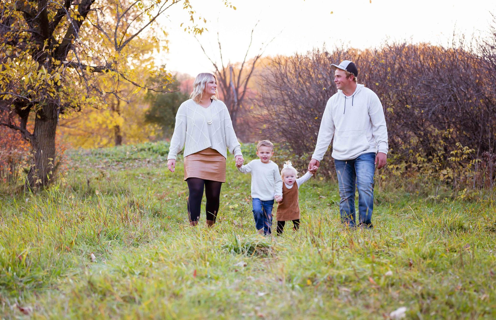 Family of four walking through a grassy field, holding hands, with autumn foliage in the background.