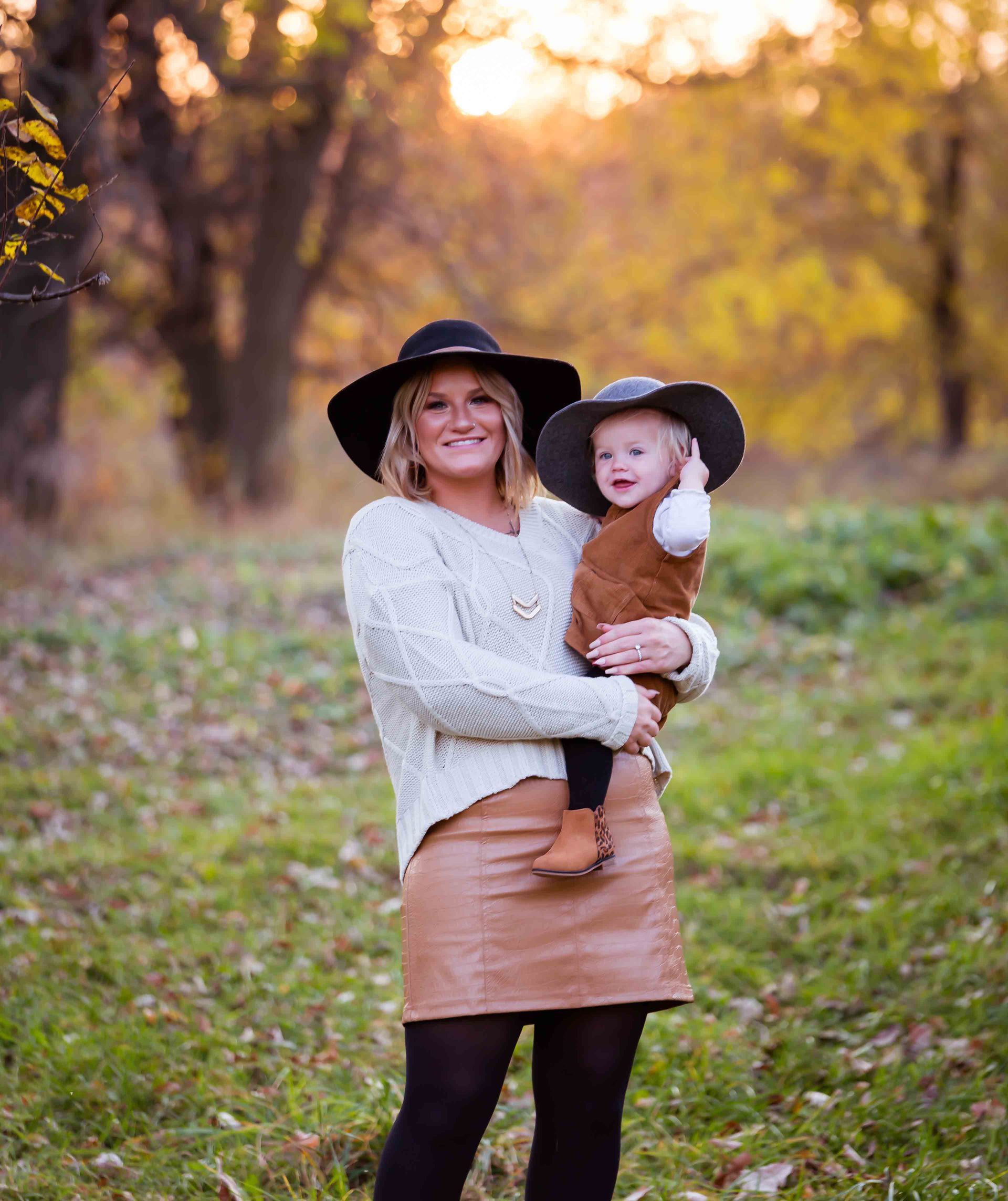 Woman holding child wearing matching hats outdoors in autumn.