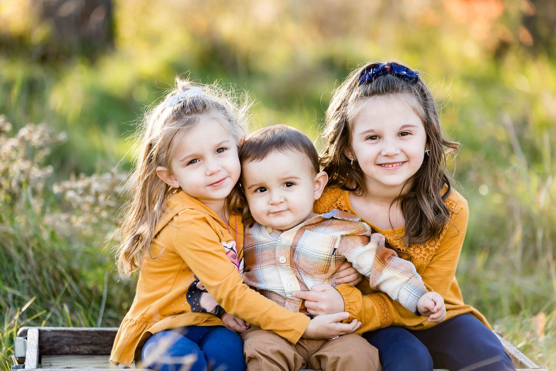 Three children, two girls in yellow hugging a boy in a plaid shirt, smiling outdoors.