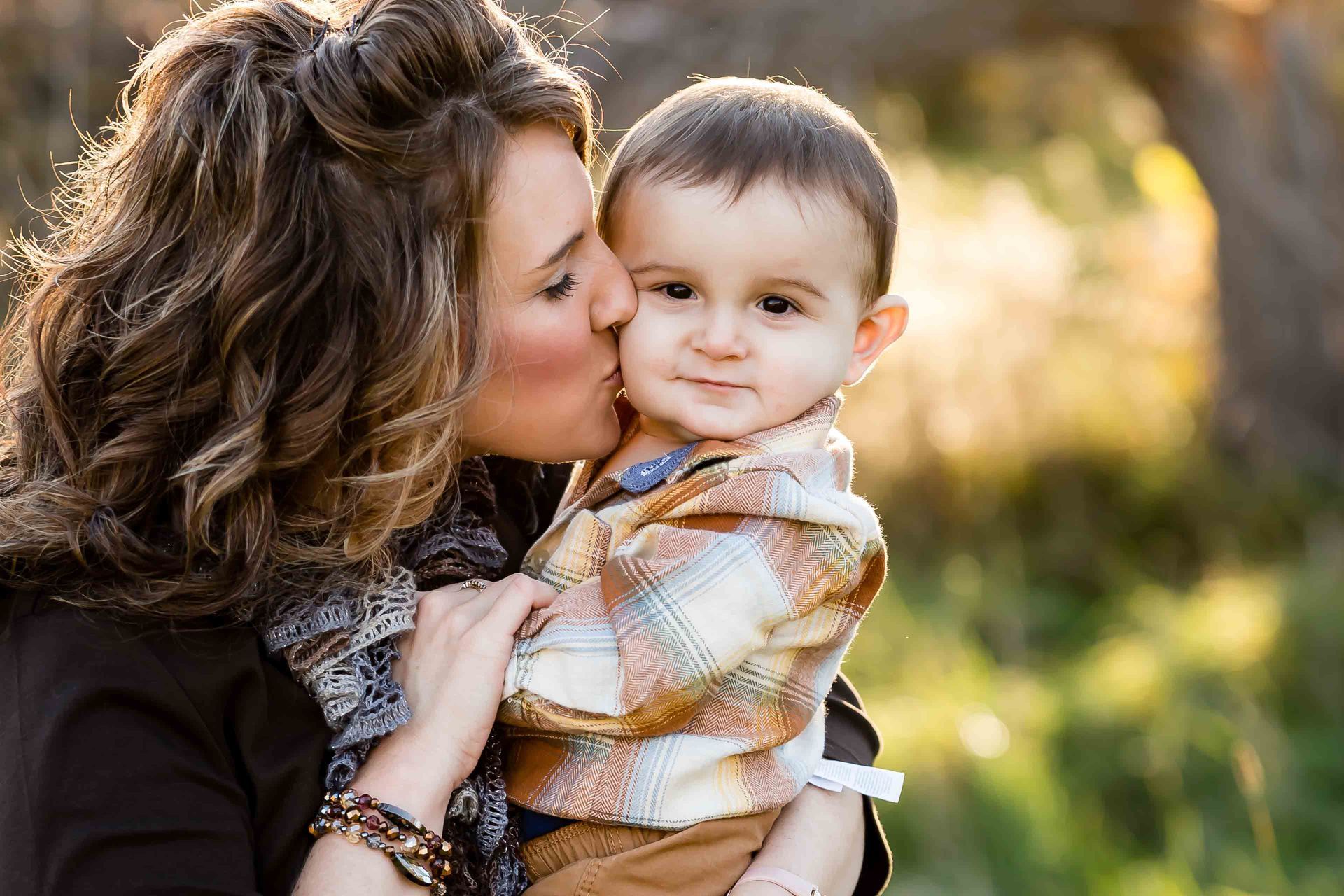 Woman kisses a child on the cheek outdoors. Both are smiling, with soft sunlight.