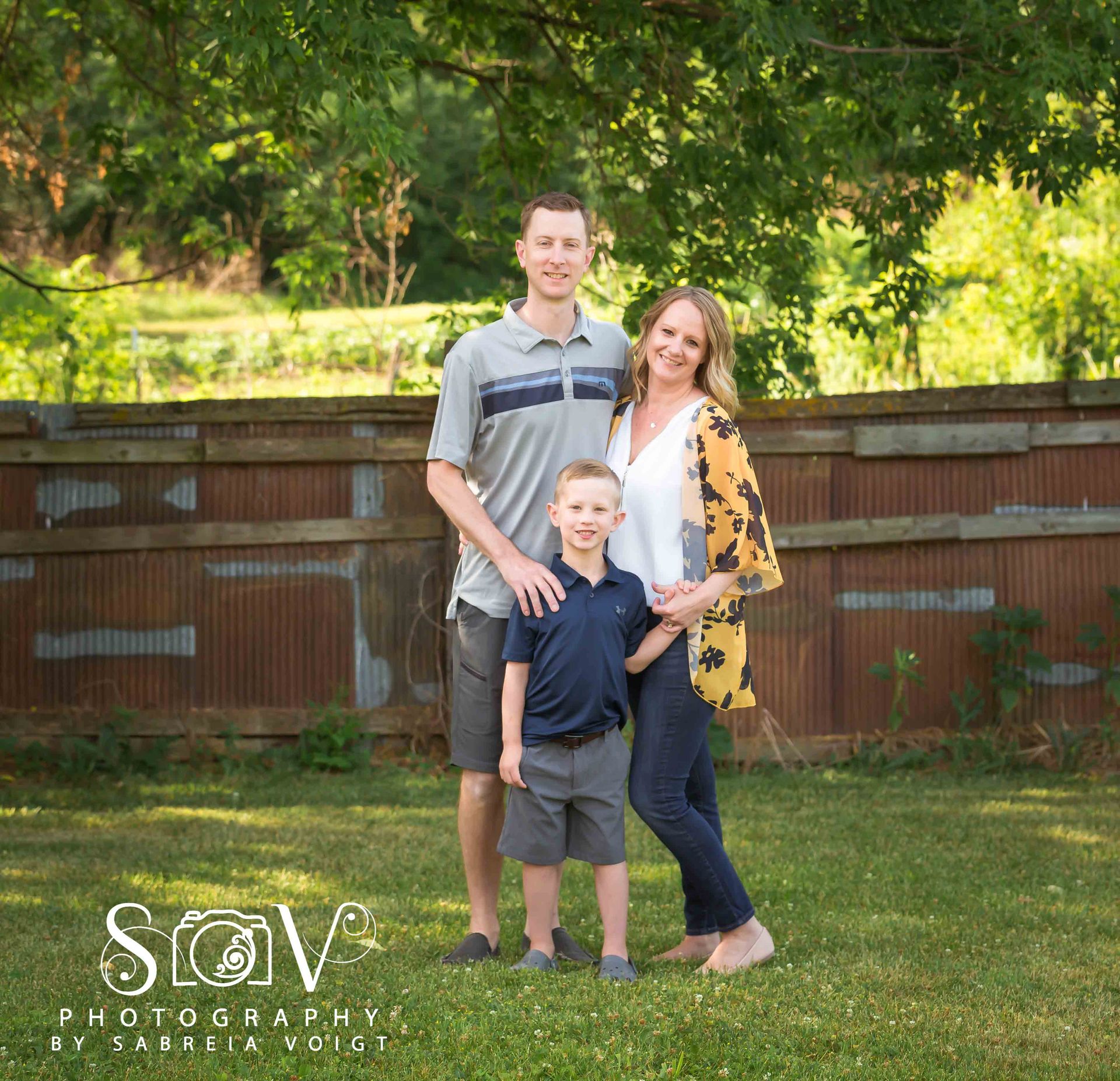 Family of three poses outdoors in front of rustic fence. Smiling people, green grass, sunny day.
