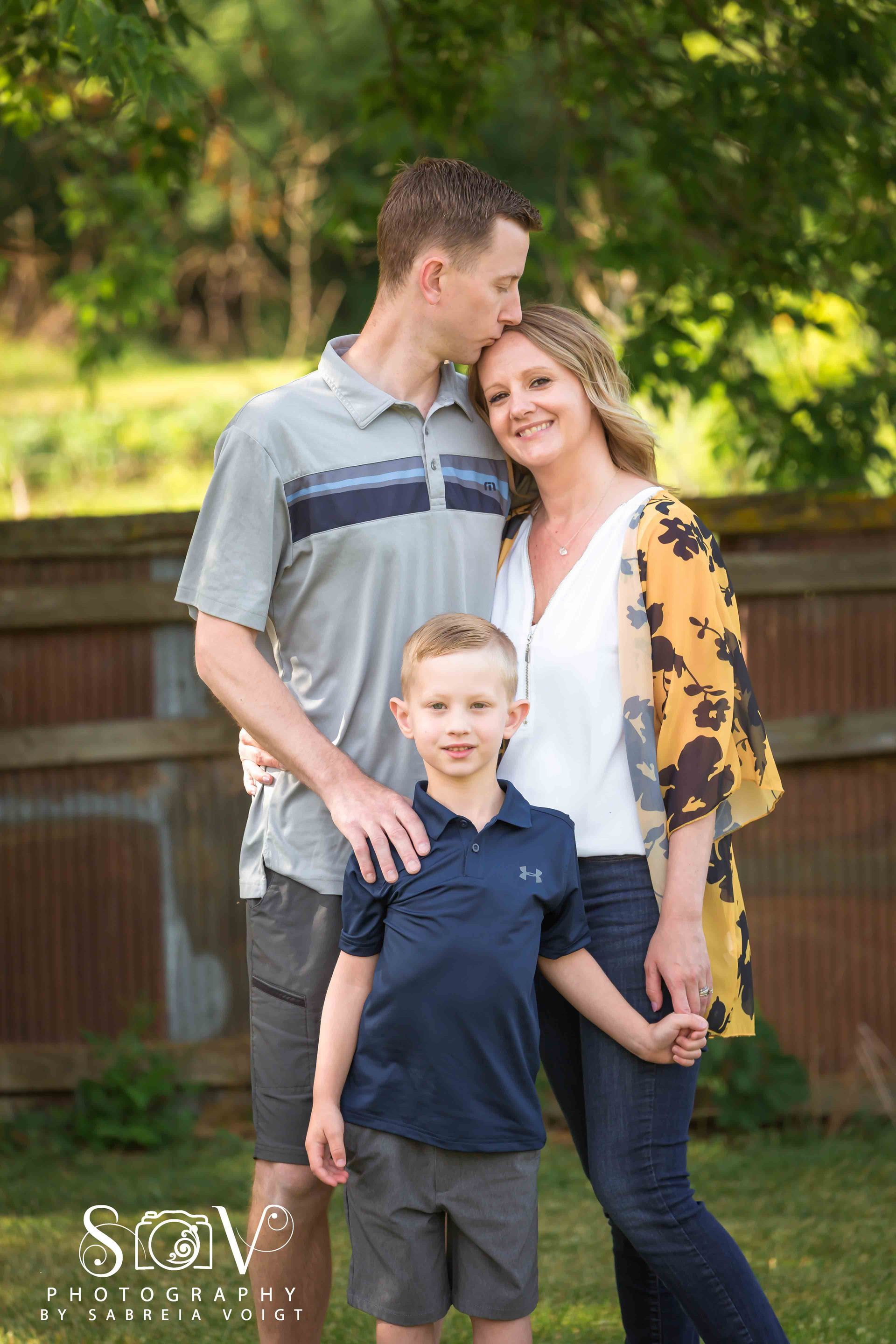 Family of three poses outdoors: man kisses woman's forehead, boy holds woman's hand, smiling.