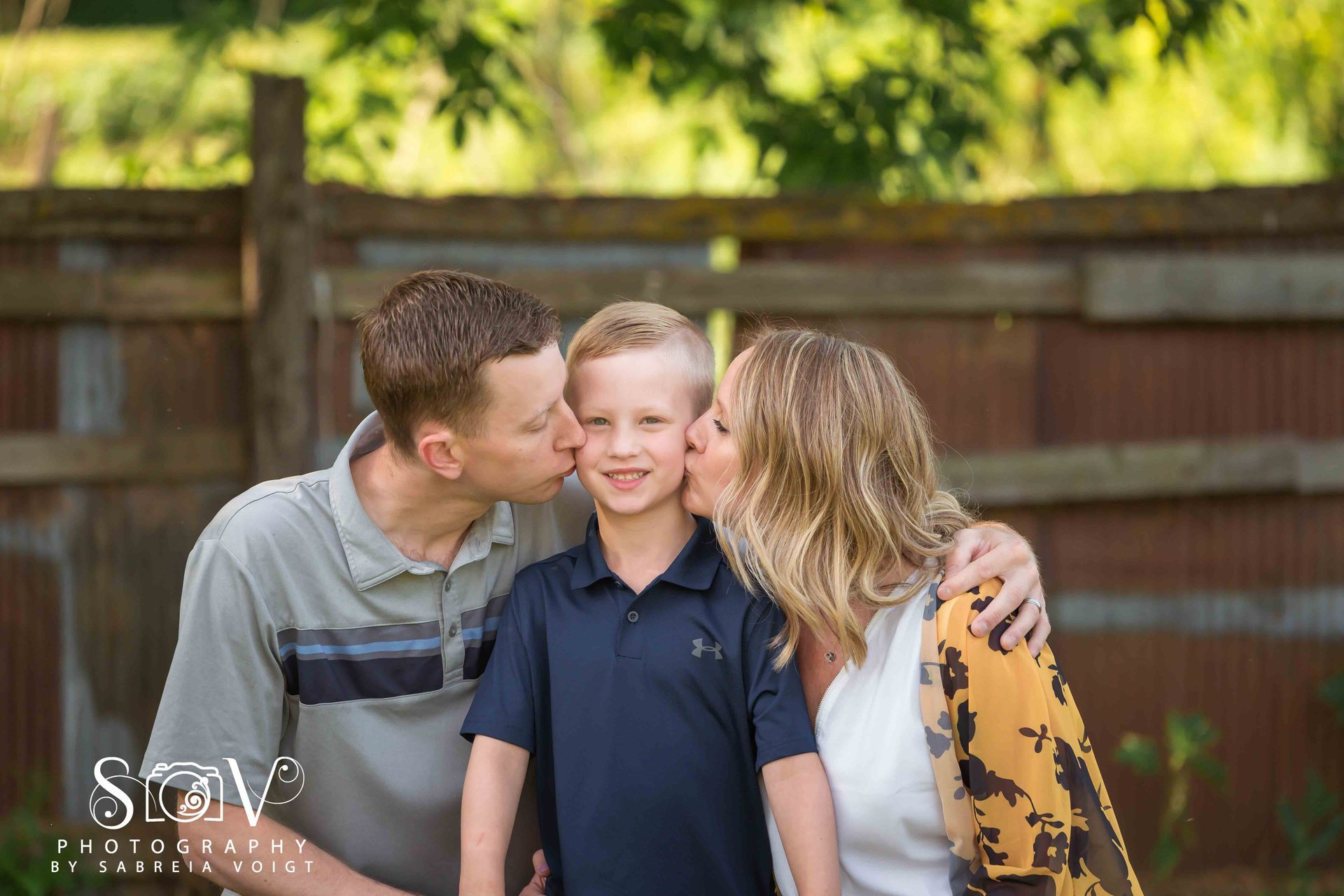 Family of three smiling while being kissed on the cheeks. Outdoors, near a fence, green foliage.