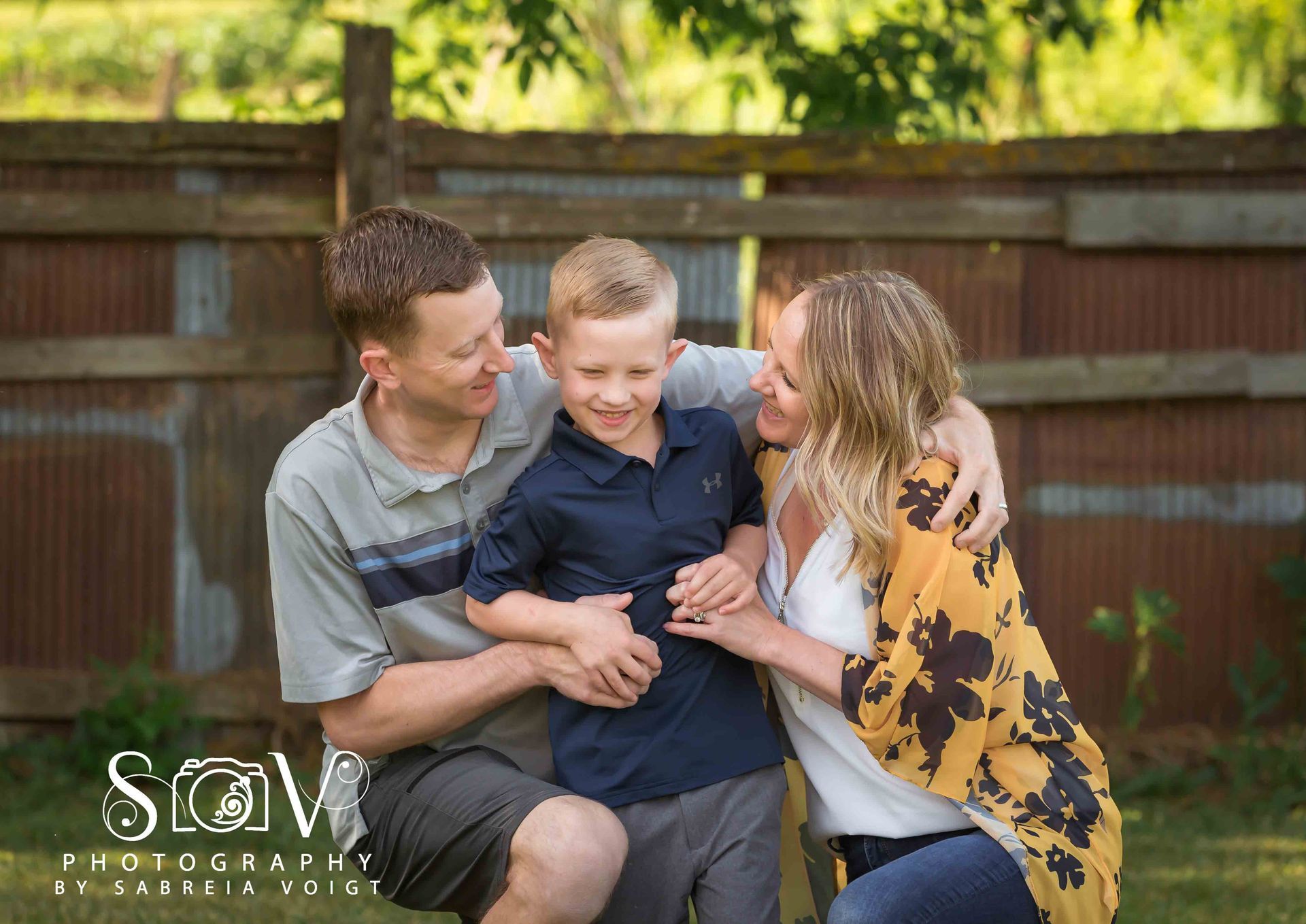 Family of three smiling, embracing outdoors; wooden fence background.