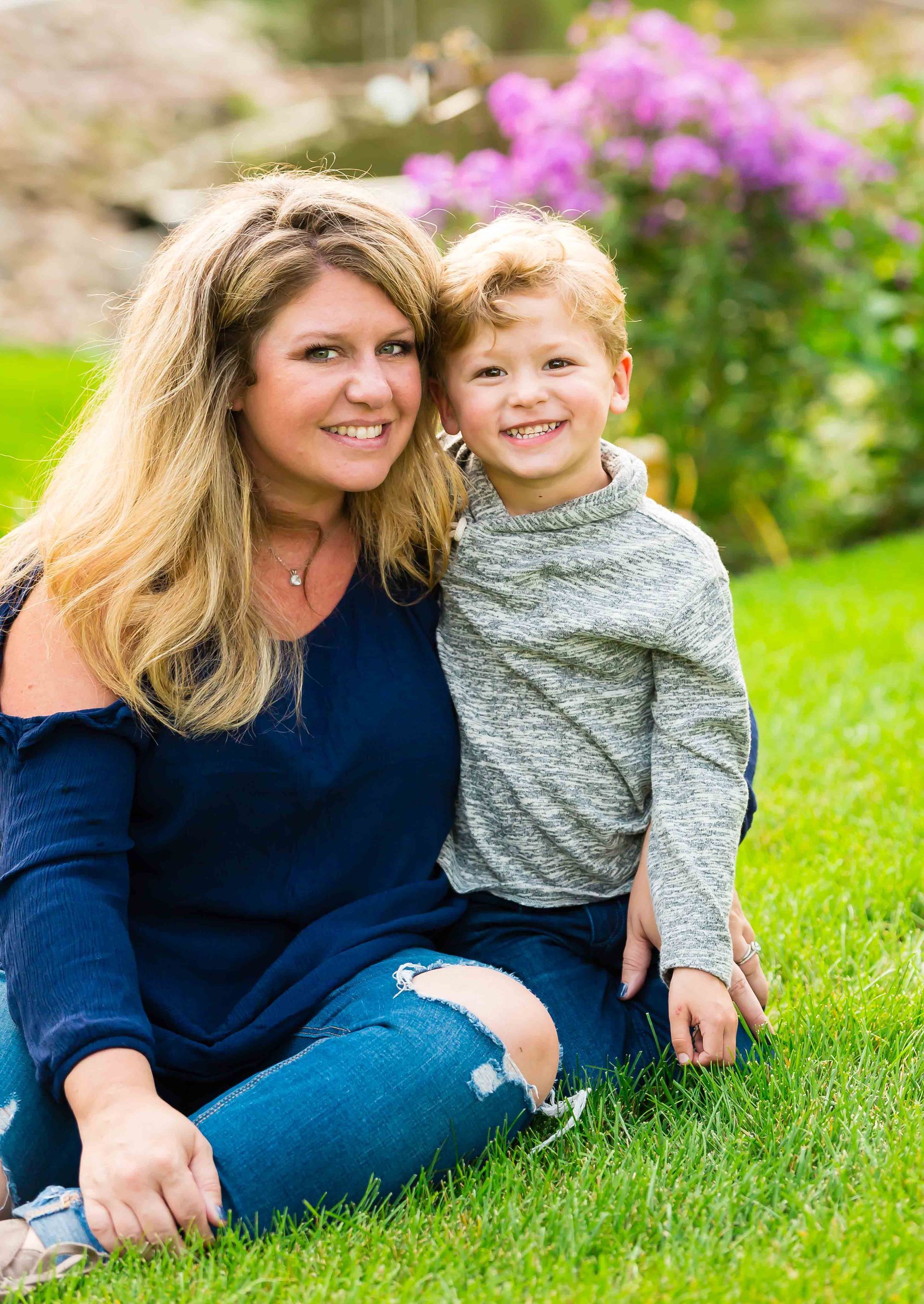 Woman and young child sitting on grass, smiling. Woman in blue shirt and ripped jeans. Child in gray sweater.